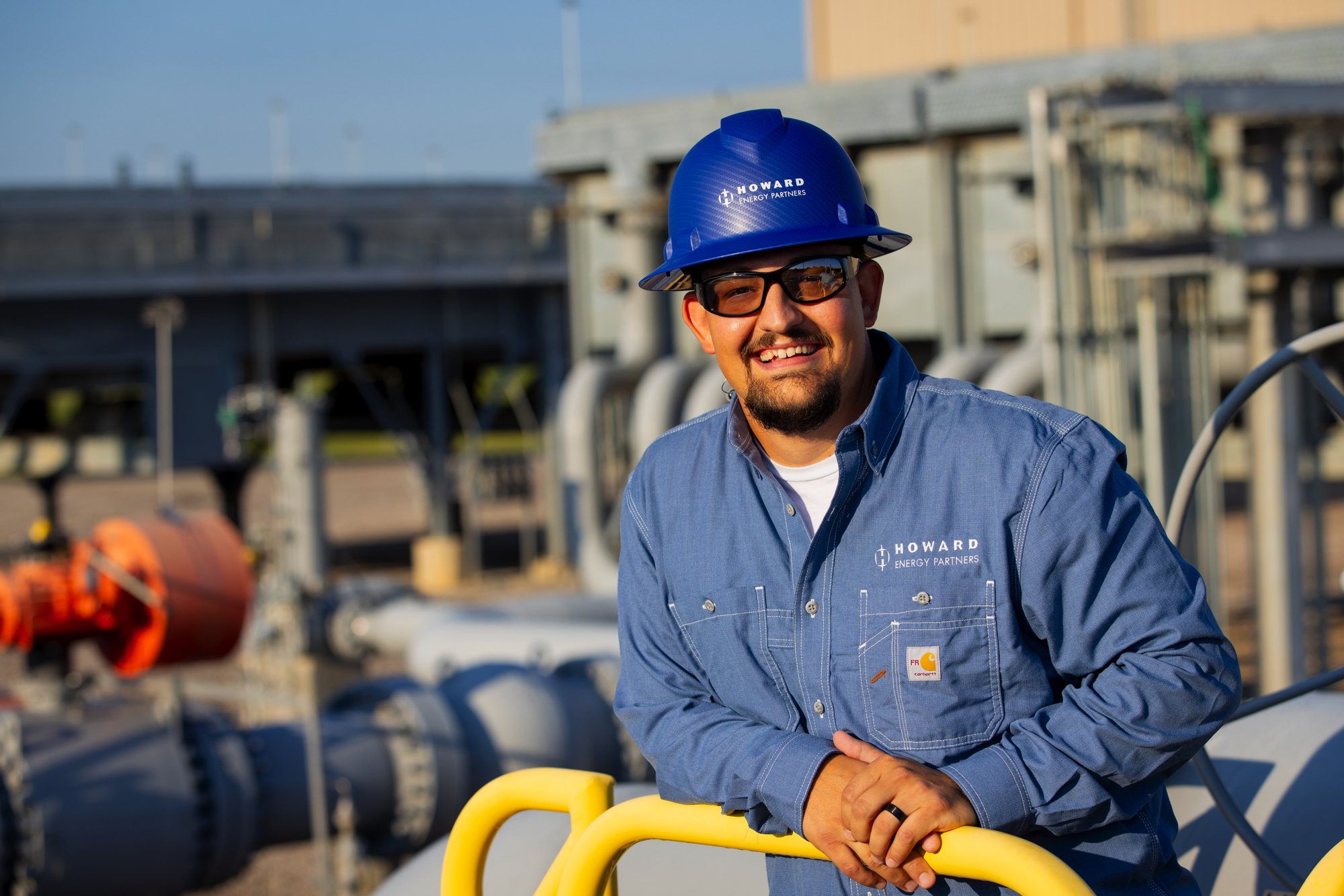 A smiling man wearing a blue hard hat and sunglasses standing outdoors on an industrial site, with large pipes and equipment in the background.