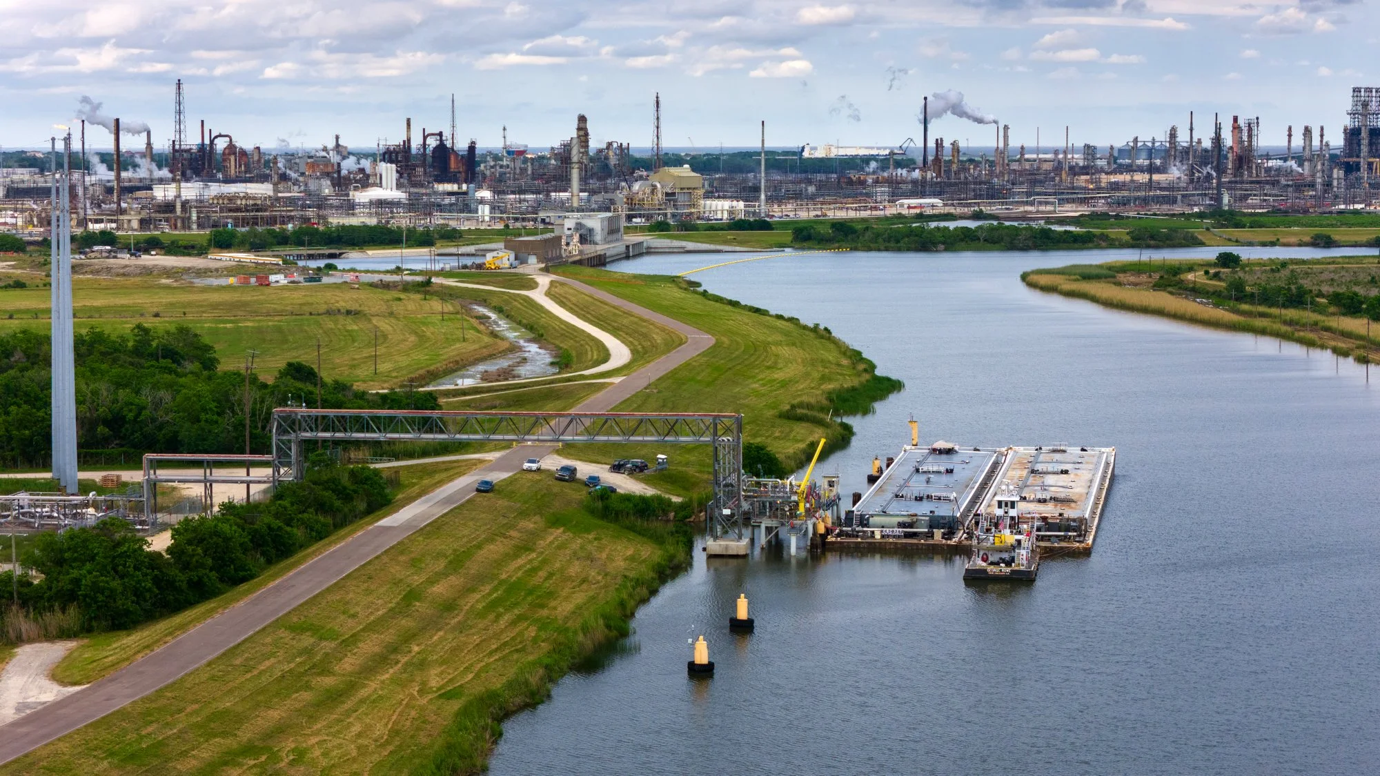 A view of an industrial area with factories and smokestacks in the background, next to a waterway with a docked barge and a small boat, surrounded by green land and a winding road.