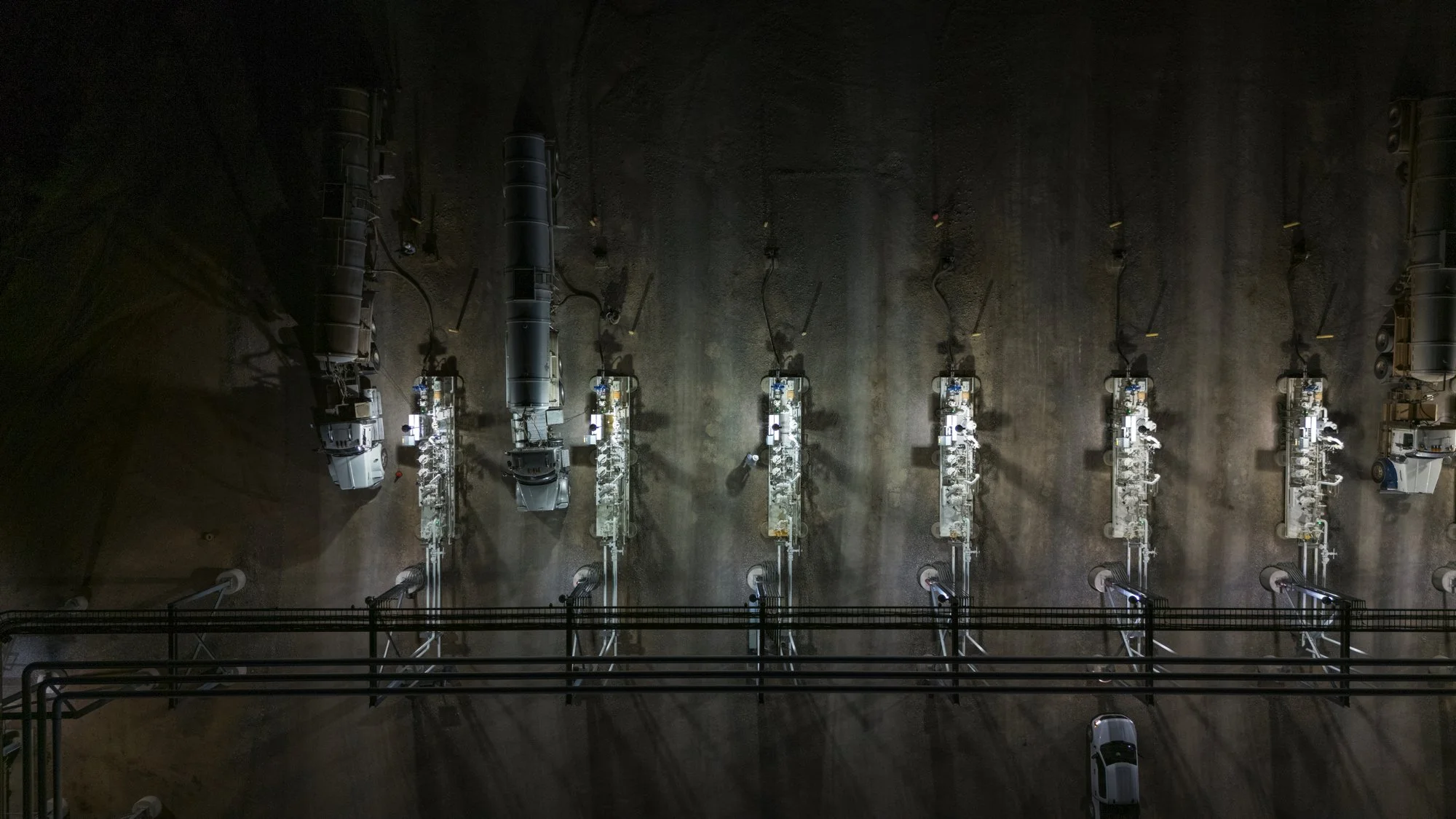 An industrial facility with multiple large tanks or reactors aligned vertically, connected by pipes and machinery, seen from above in a spacious, dimly lit room.