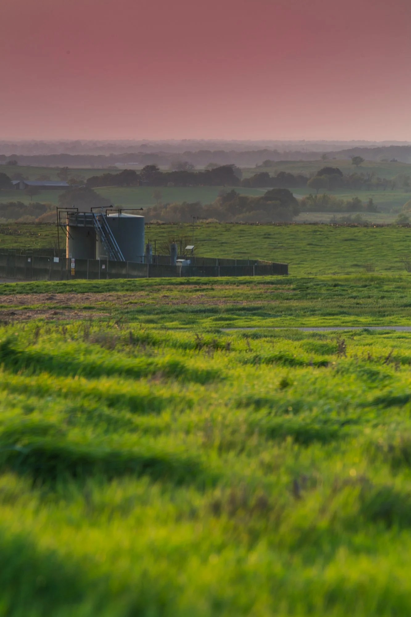 A landscape with green fields and rolling hills in the distance, with a blue industrial water tank near a fence, under a pinkish sky at sunset.