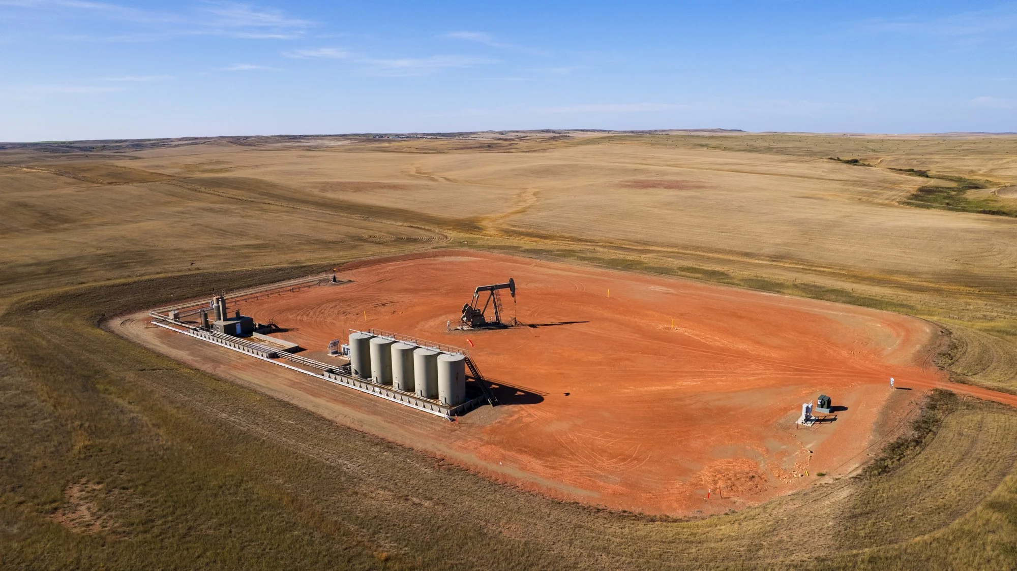 An oil drilling rig with four storage tanks and support structures on dirt land in an open, flat landscape with distant fields and a blue sky.