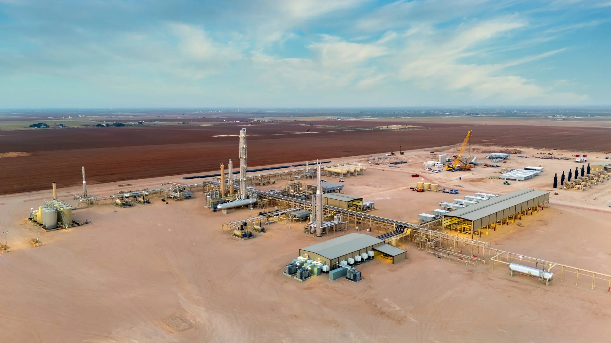 Aerial view of an oil drilling and production site with multiple equipment, storage tanks, and a crane, set in a desert landscape with agricultural fields in the background.