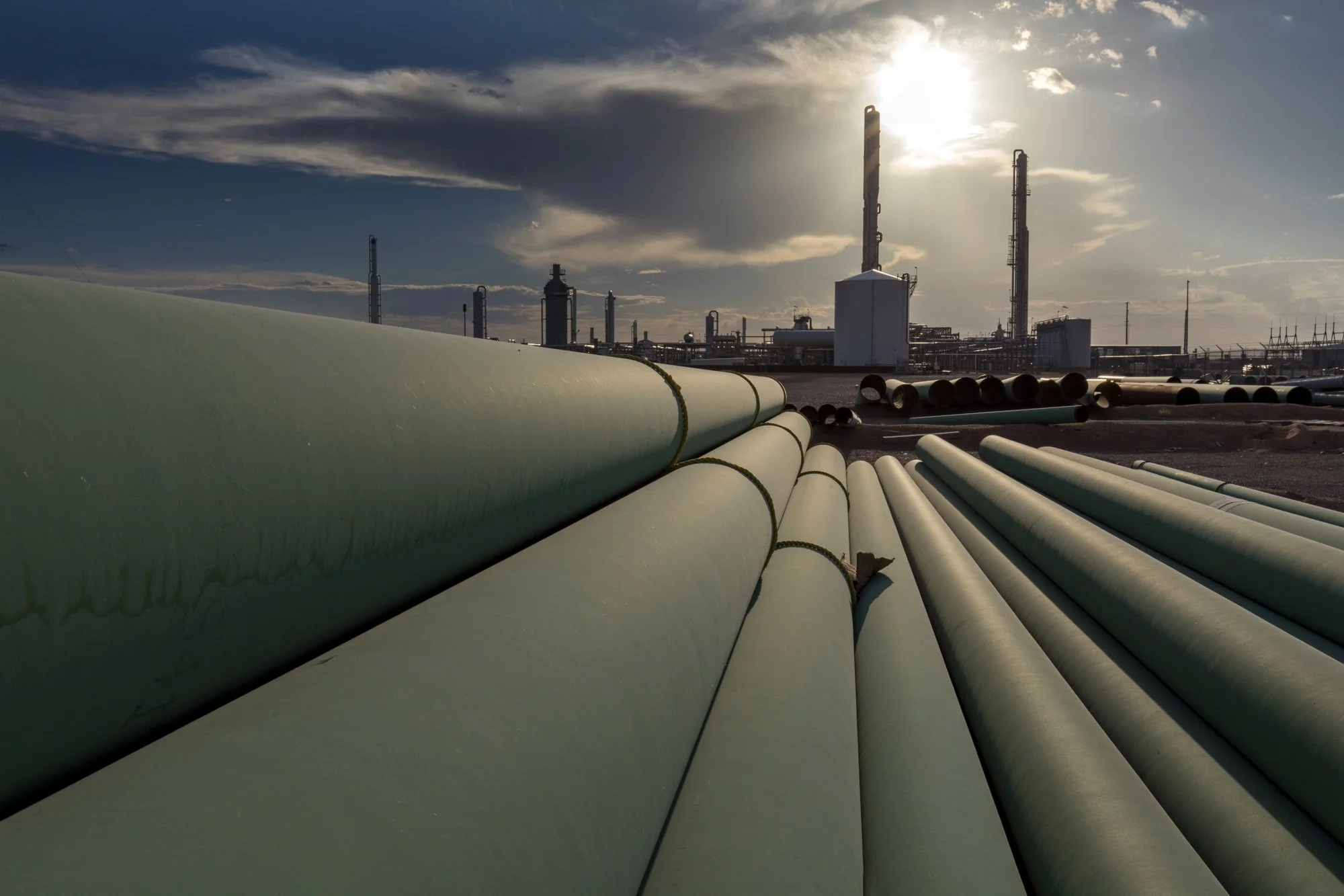 Large light green pipes laid on the ground leading to an industrial refinery with tall structures and towers under a partly cloudy sky during sunset.