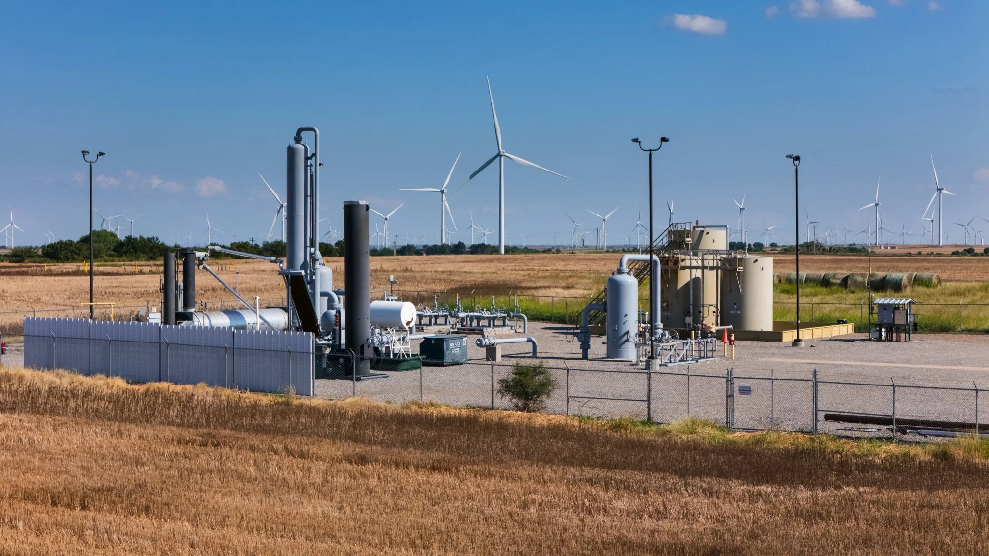 An industrial facility with pipes and storage tanks in a field, with wind turbines in the background under a blue sky.