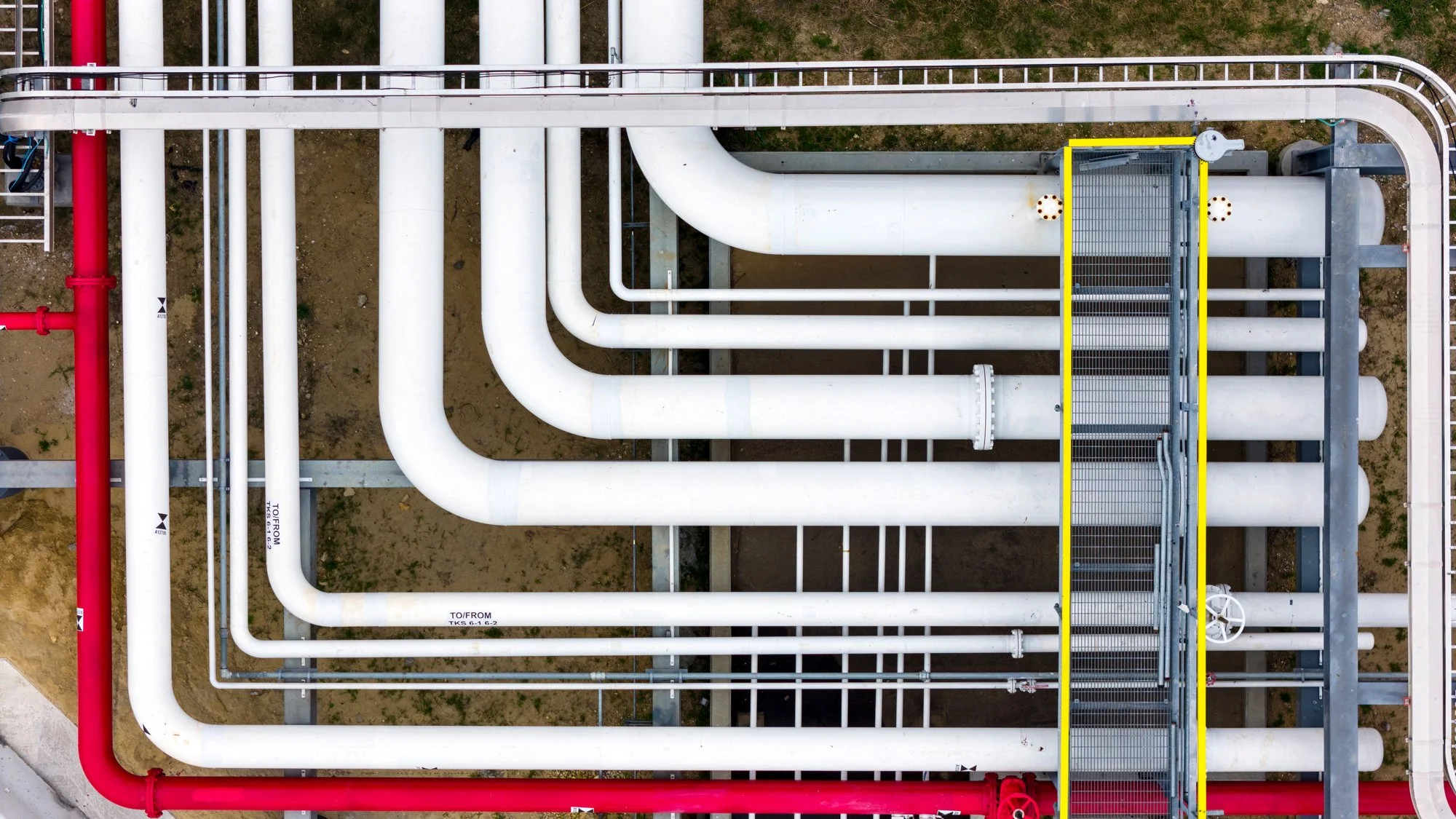 An overhead view of industrial pipes, with large white pipes and a few smaller ones on a concrete and dirt surface, including a yellow-railed metal platform.