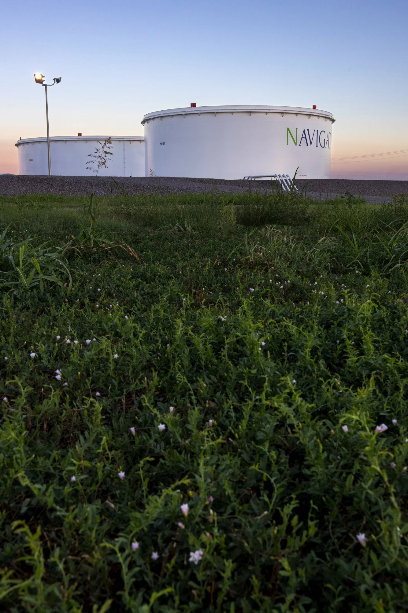 Large white fuel storage tanks with the word 'NAVIGANT' on them, set against a clear sky at sunset, with green vegetation in the foreground.