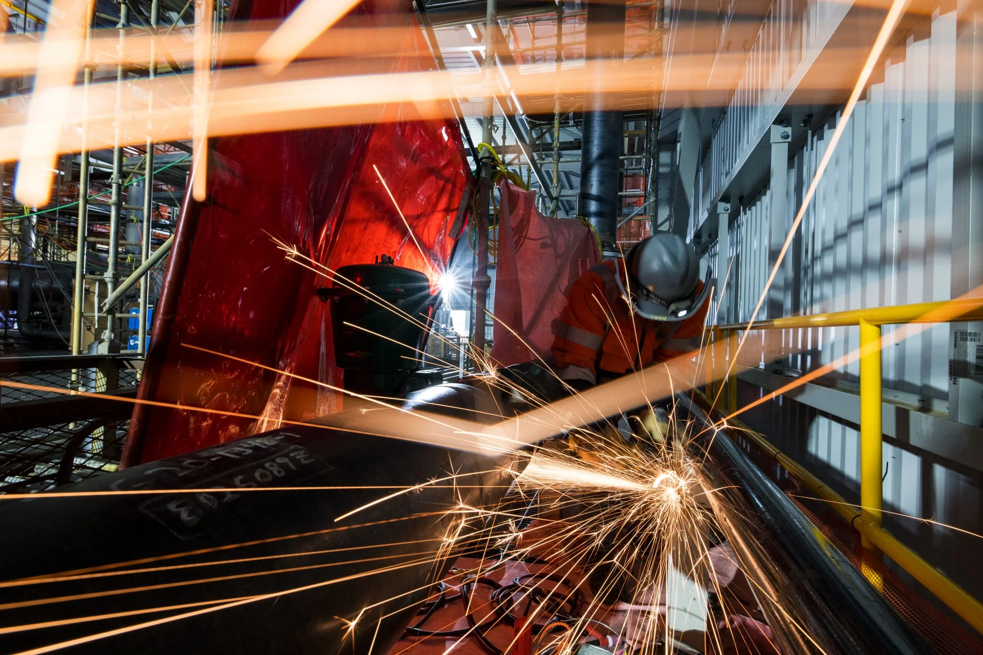 Worker welding a pipe on an industrial construction site with sparks flying.