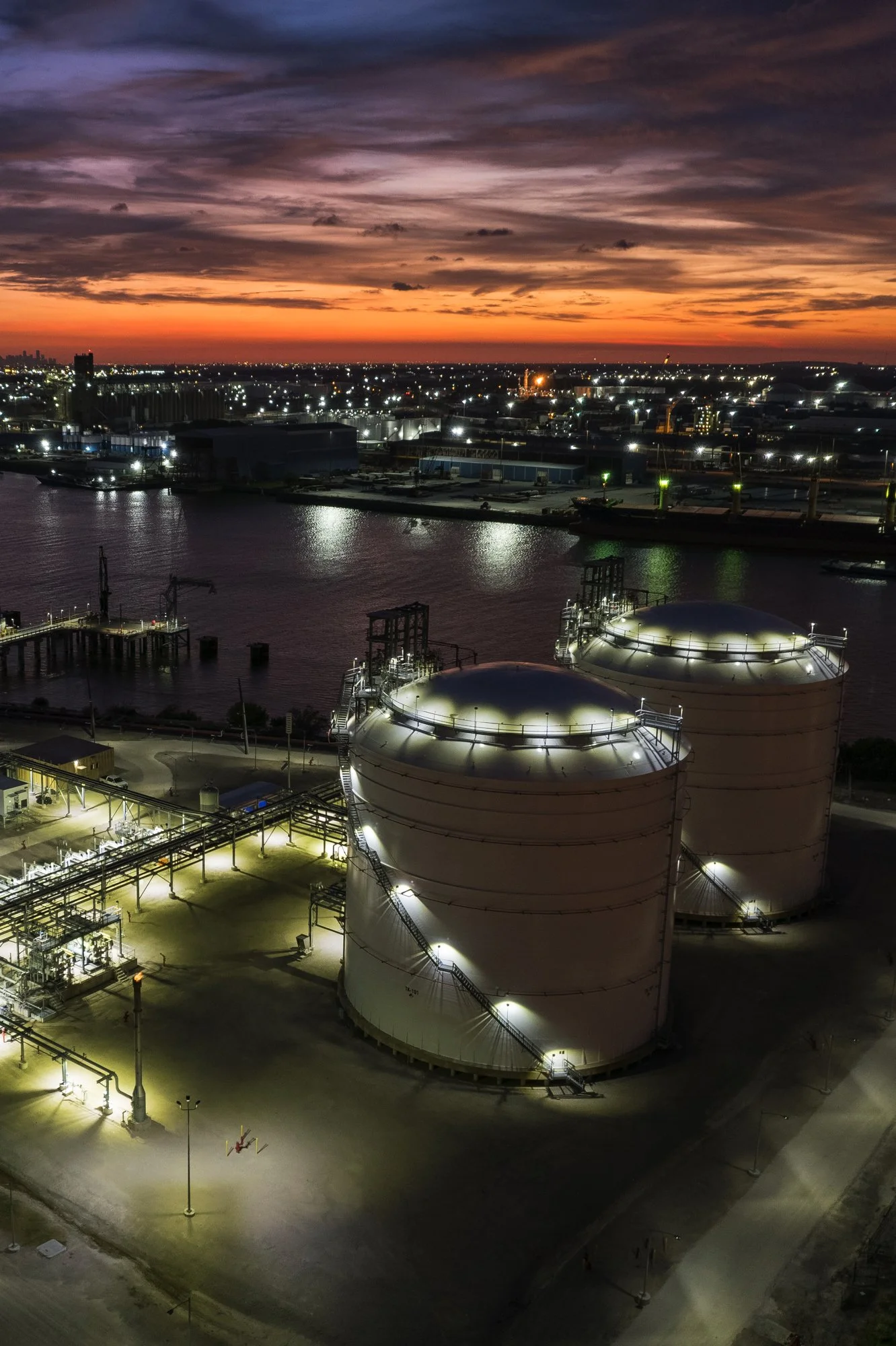 Nighttime view of an industrial facility with two large white storage tanks illuminated, near a body of water, and a city skyline with sunset sky in the background.