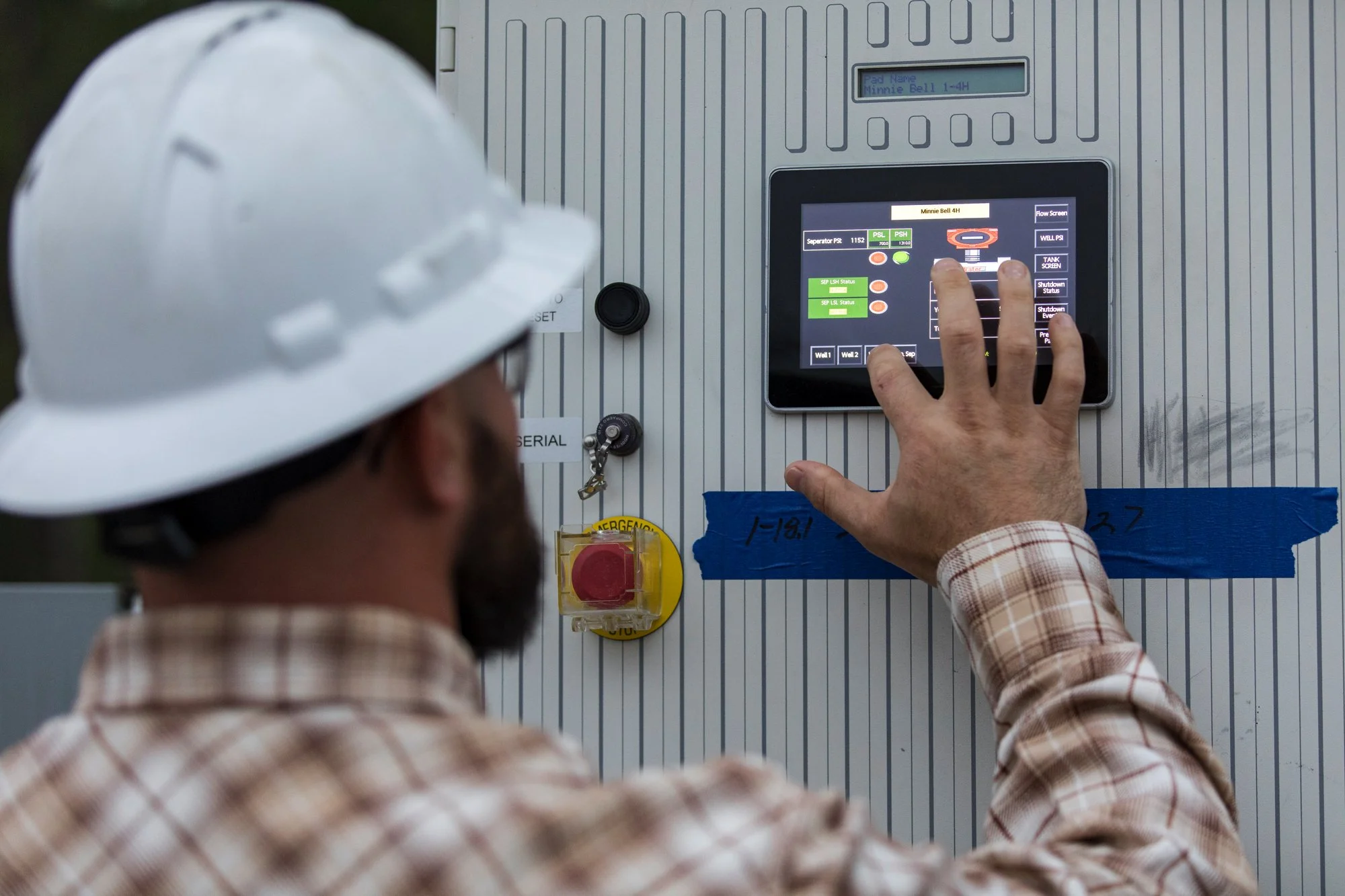 A male worker wearing a white safety helmet and plaid shirt operating a control panel with a touchscreen interface, mounted on a wall with a yellow emergency stop button nearby.