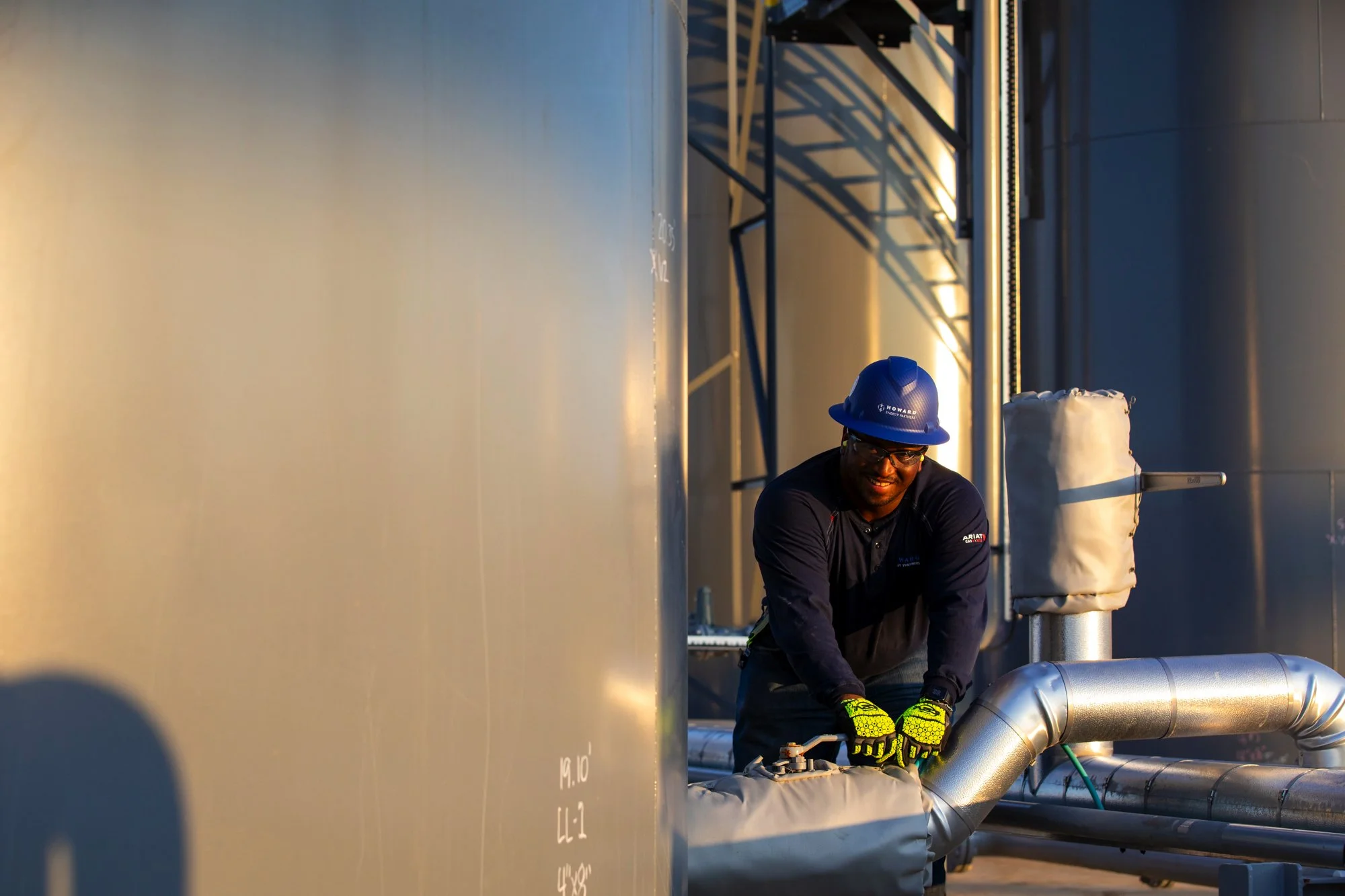 A worker wearing a blue safety helmet, glasses, black long-sleeve shirt, and yellow gloves is smiling as he works on metallic pipes outdoors near industrial tanks or silos.
