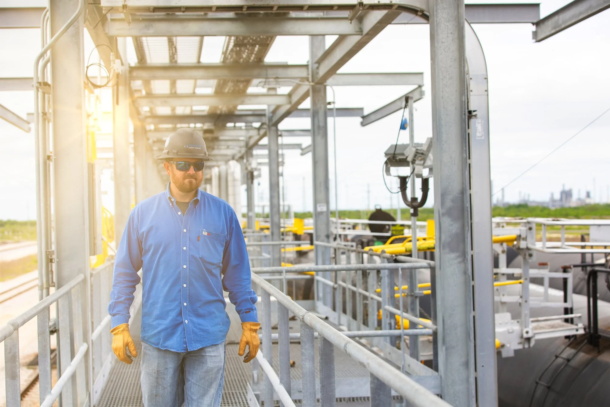 A man in a blue shirt, safety glasses, gloves, and a safety helmet walking along an industrial platform at a solar energy plant. Solar panels and equipment are visible in the background.
