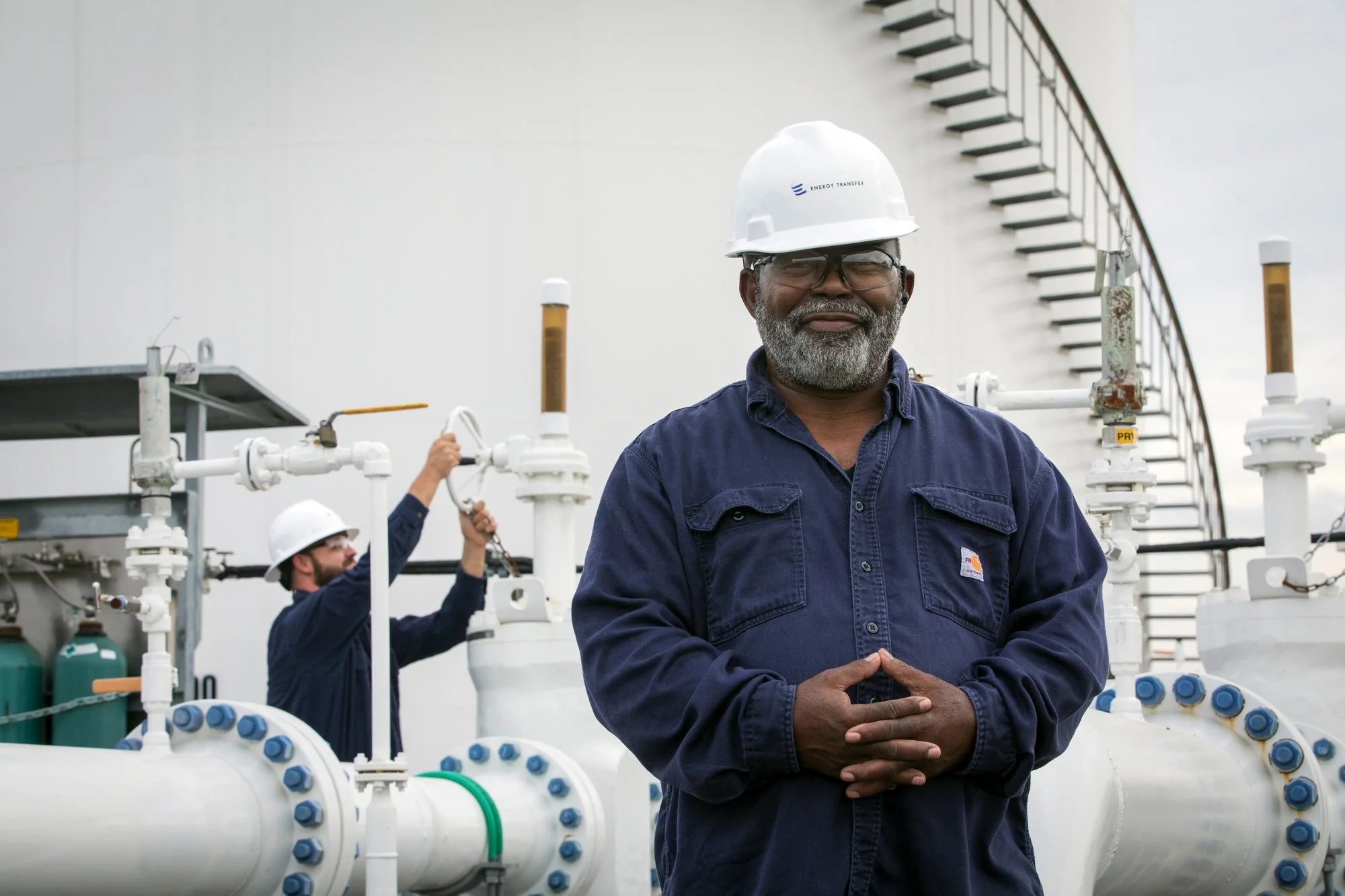 A smiling man wearing a white safety helmet and glasses standing in front of industrial equipment. In the background, another worker is adjusting pipes on a large industrial facility.