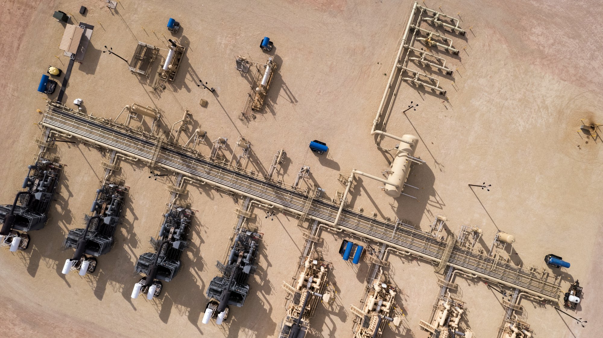 Aerial view of an industrial site with pipelines, large machinery, storage tanks, and blue tanks on a beige dirt ground.