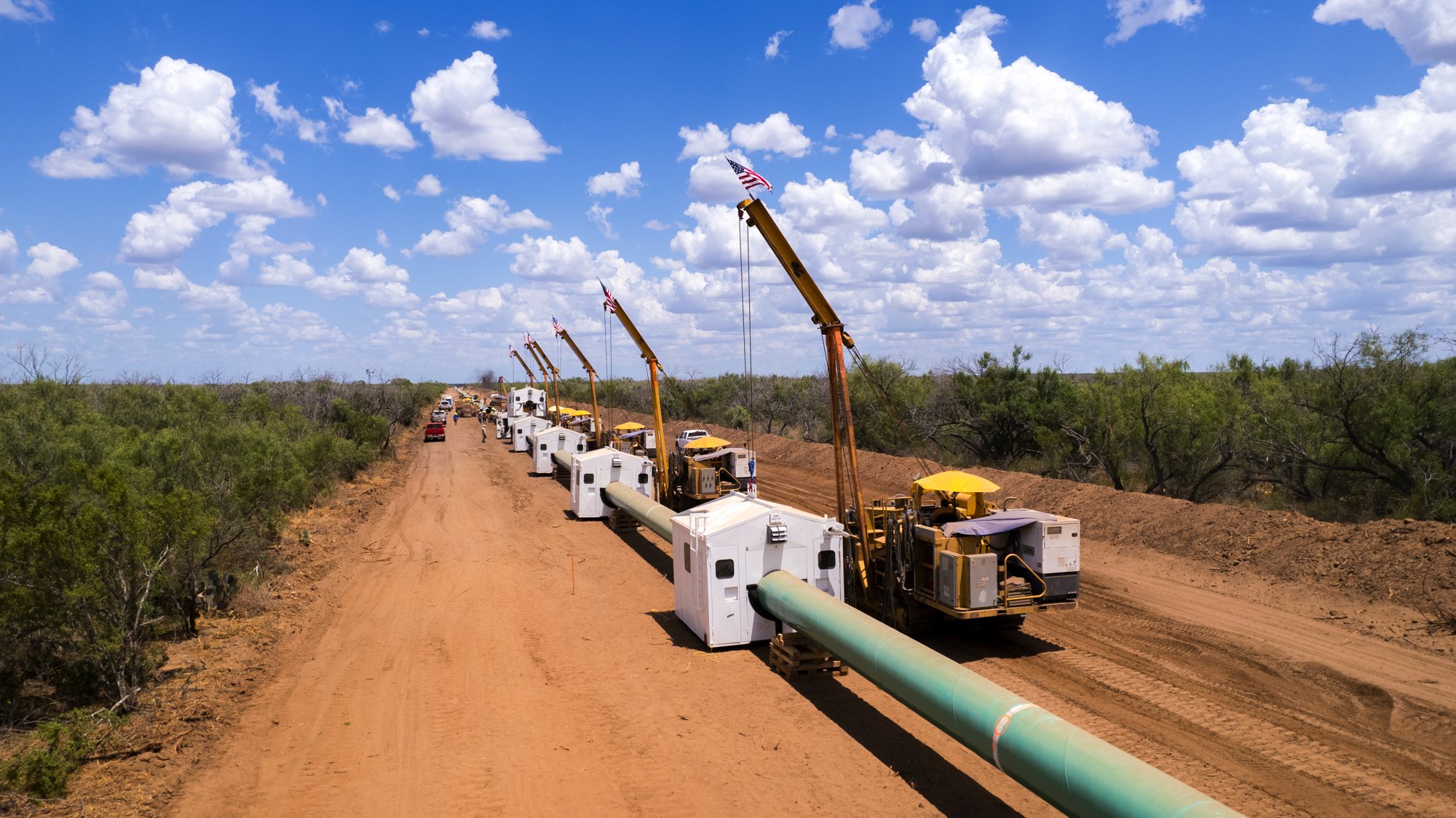Construction workers and equipment install a pipeline along a dirt road in a rural area with scattered trees under a blue sky with clouds.