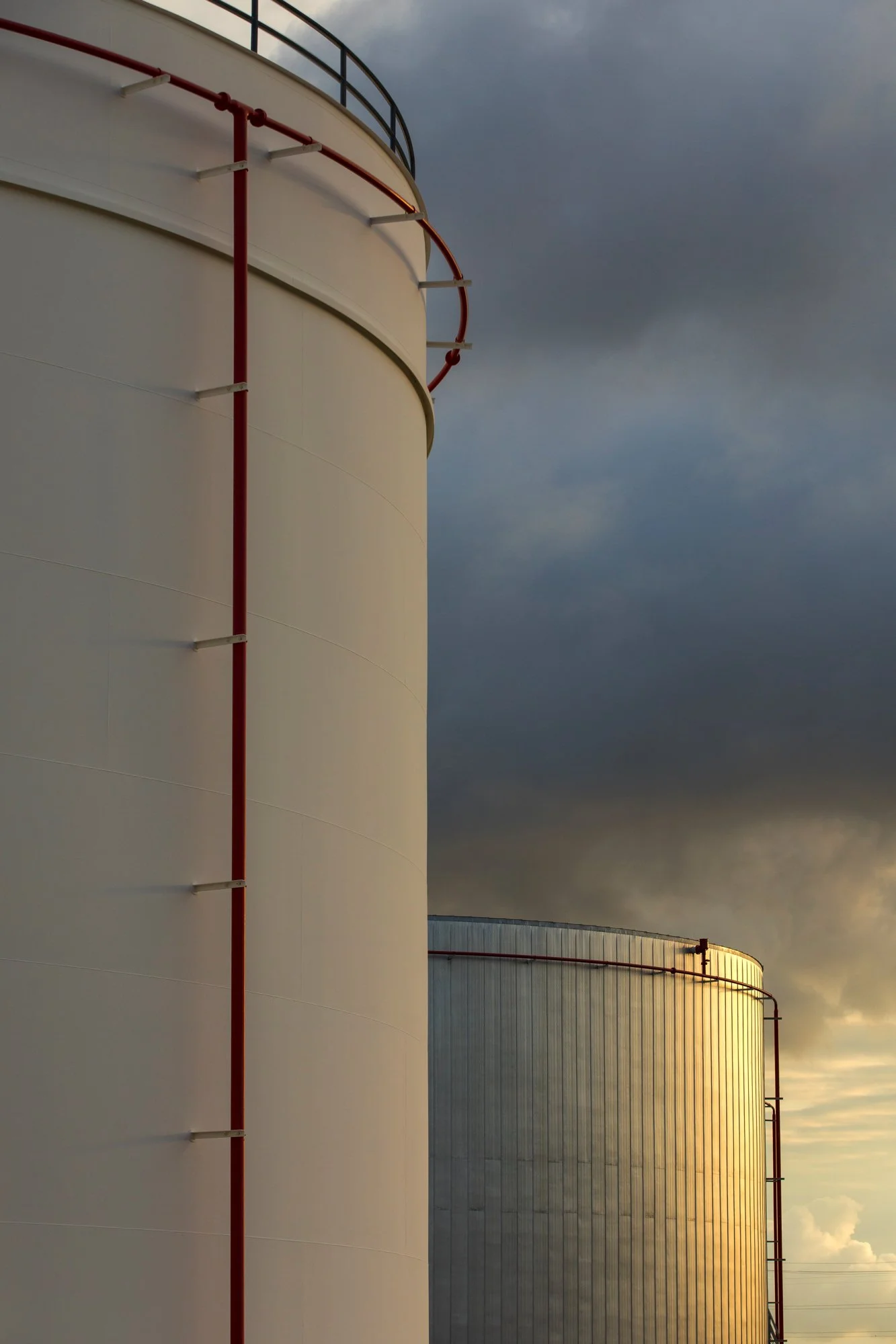 Close-up of large industrial storage tanks with red piping and a cloudy sky in the background.