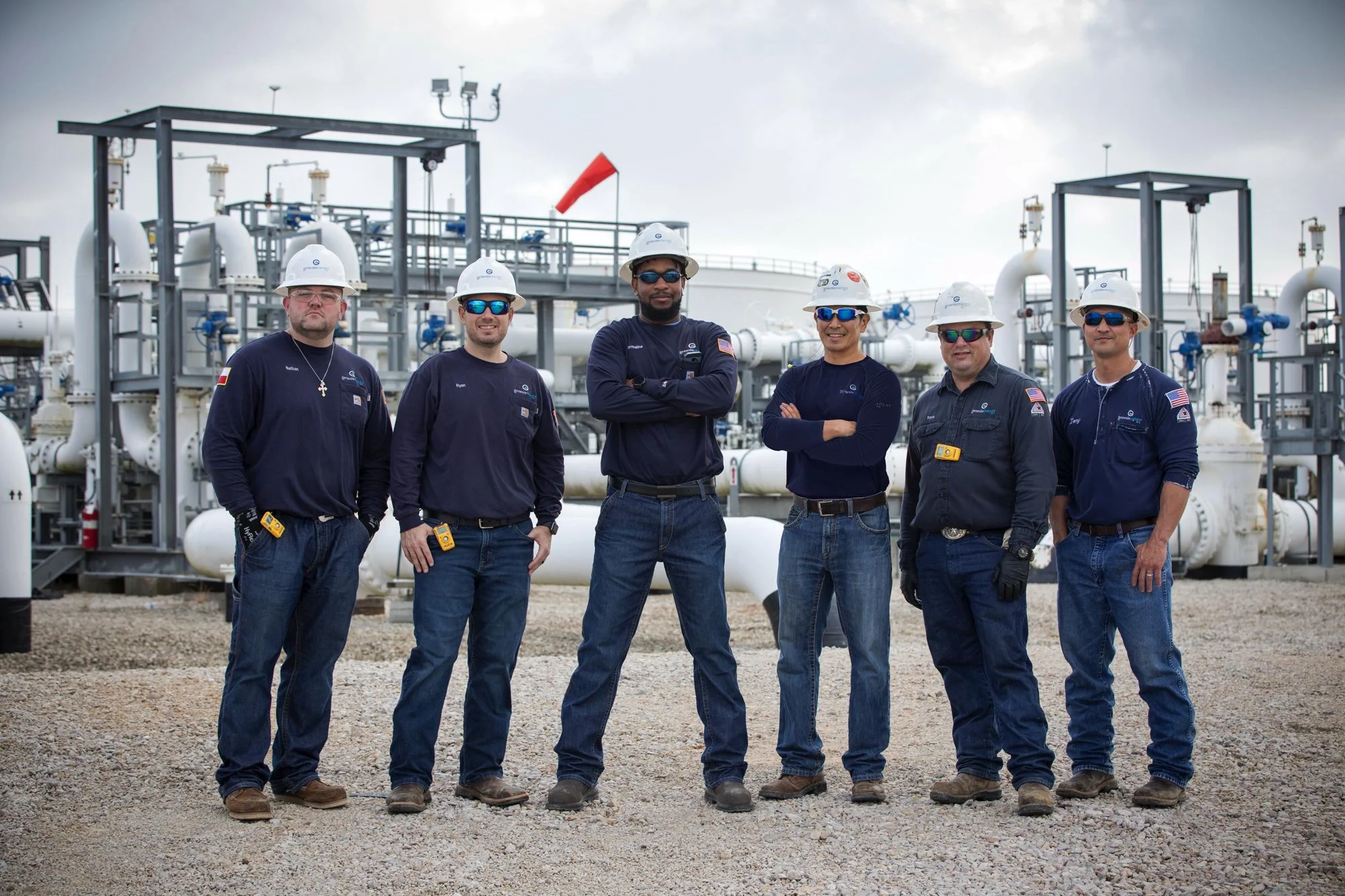 Group of six workers standing in front of industrial equipment at an oil or gas facility. They are wearing protective helmets, sunglasses, and work uniforms.