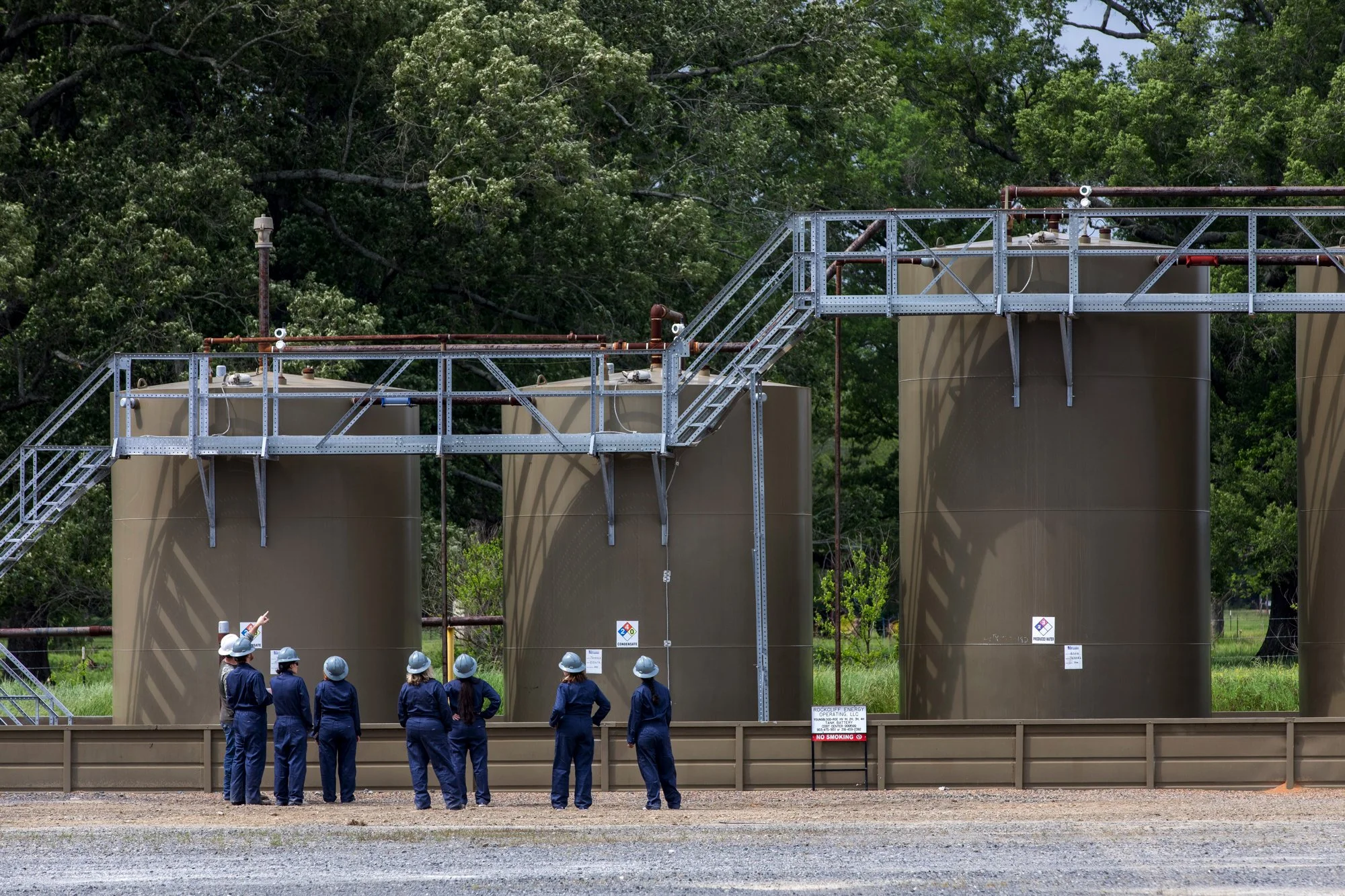 A group of people wearing blue coveralls and safety helmets, standing in front of large industrial storage tanks with piping and metal walkways, with a wooded area in the background.