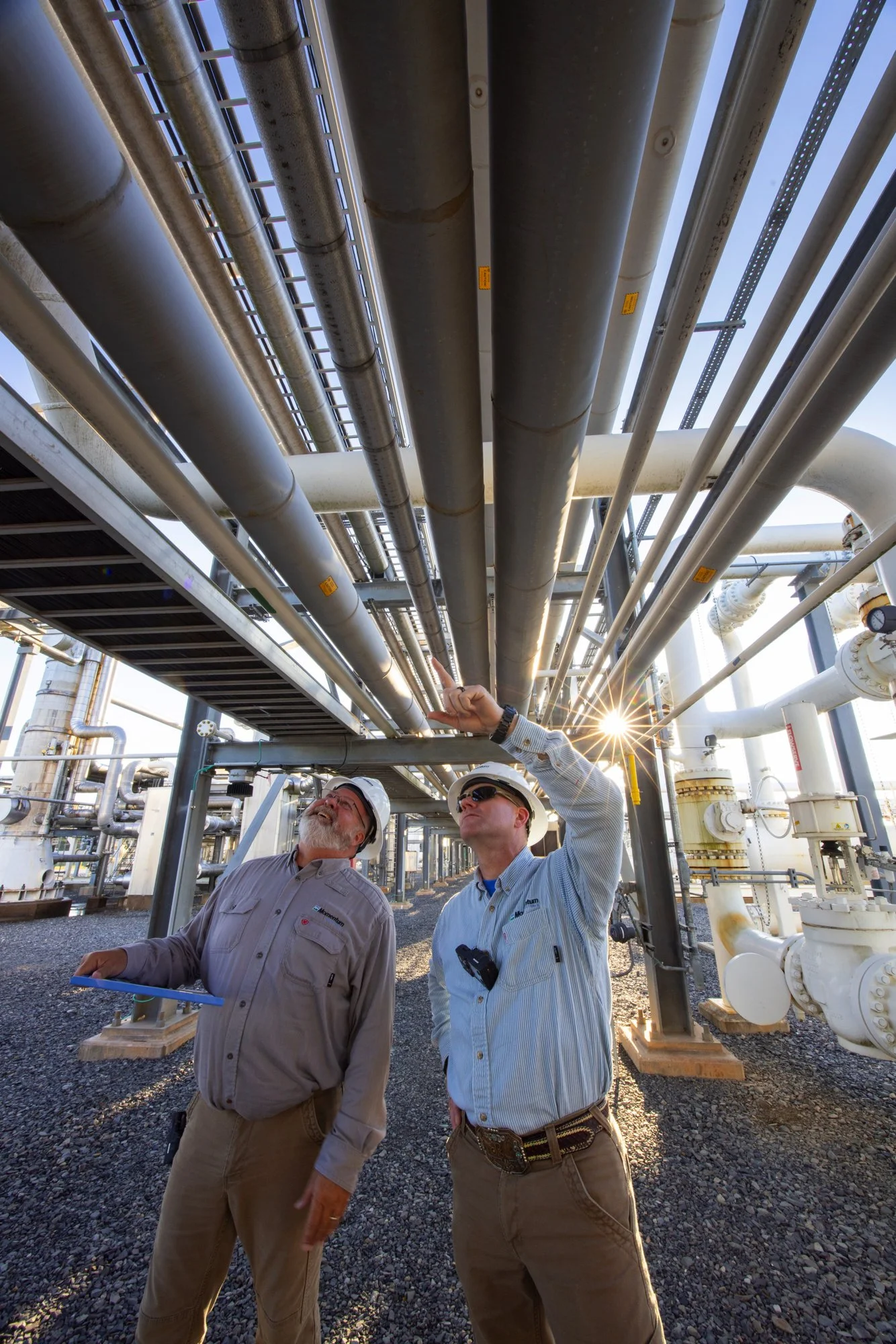 Two engineers in safety helmets inspecting industrial pipes and equipment in an outdoor industrial site, with sunlight shining through the pipes.