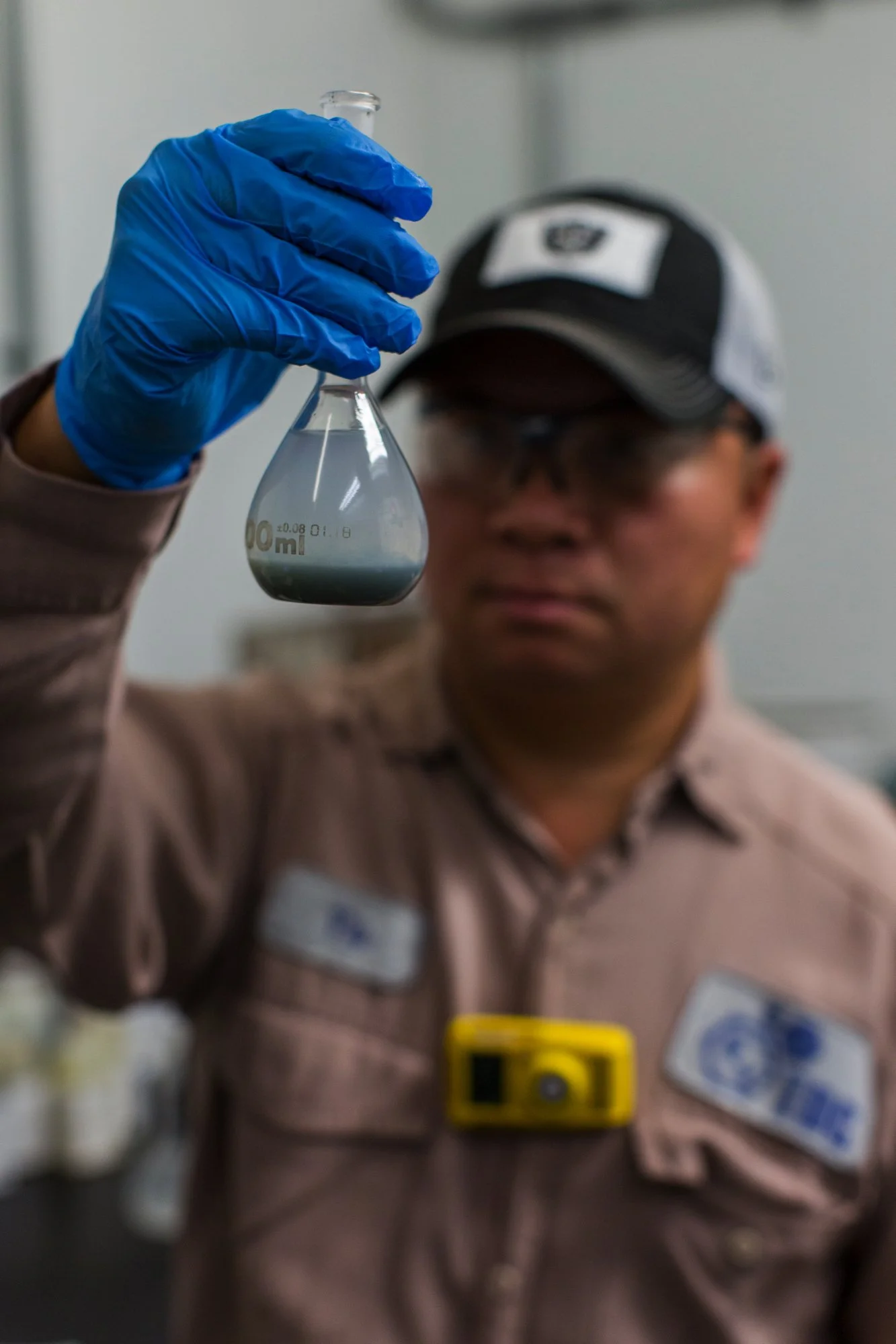 A man wearing protective glasses, a cap, and a tan uniform with badges is holding a glass Erlenmeyer flask containing a cloudy, grayish liquid, using a blue glove.