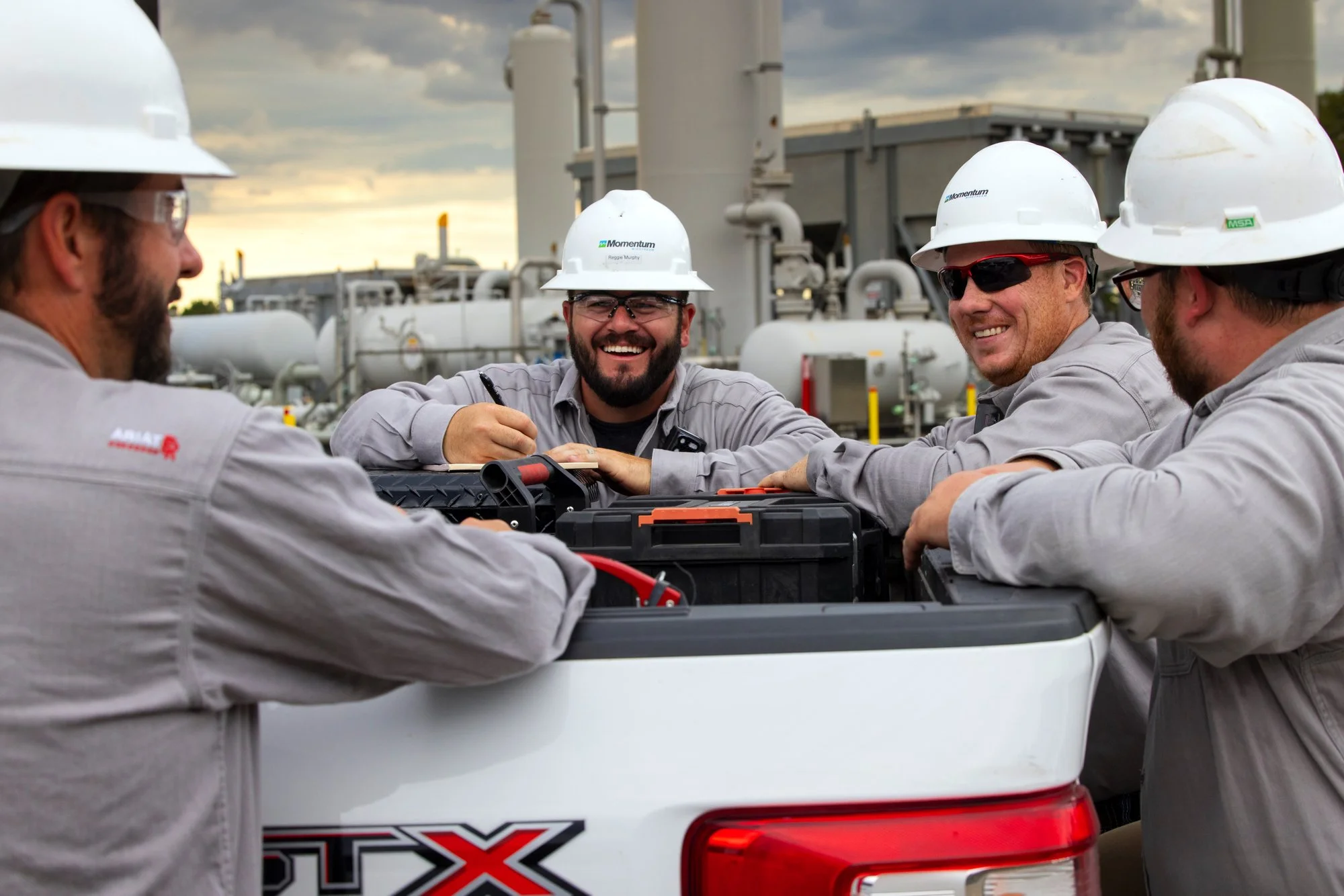Four workers wearing safety gear, including hard hats and safety glasses, gather around a toolbox or equipment on the back of a truck at an industrial site during sunset.