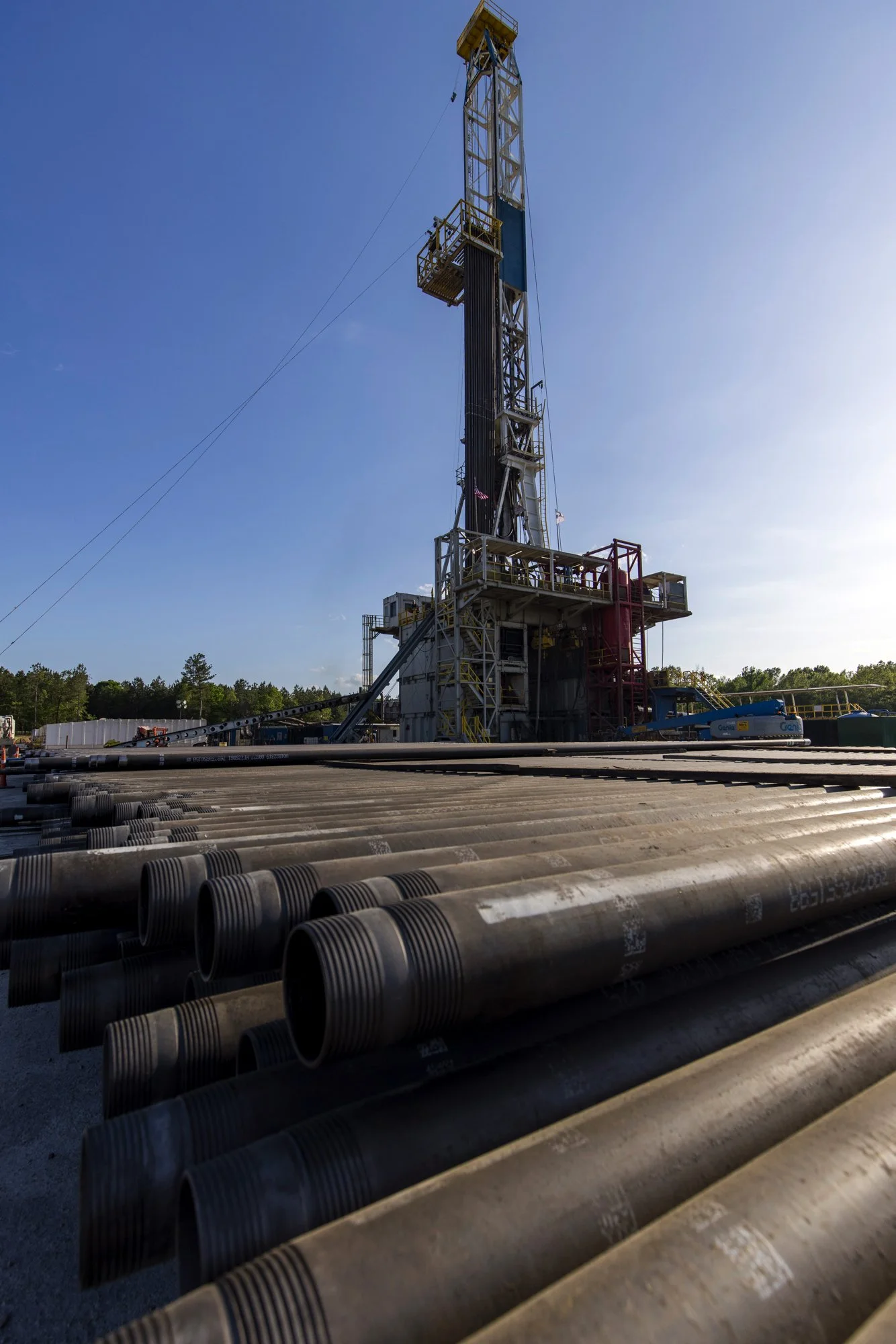 Oil drilling rig with pipes in the foreground, set against a sunny sky with trees in the background.