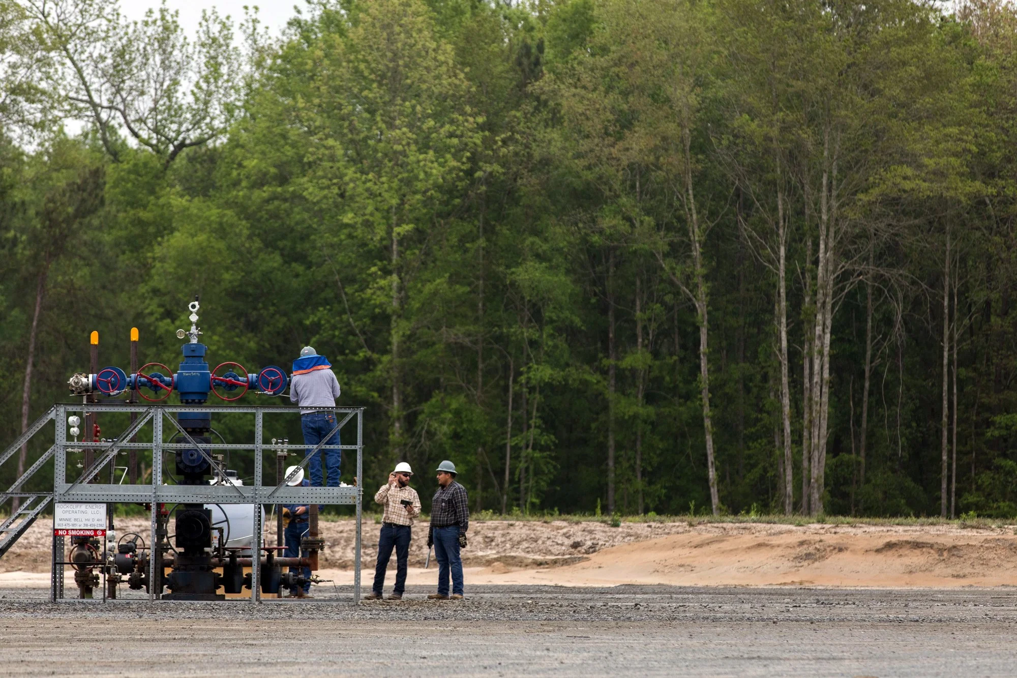Group of four workers at a natural gas or oil drilling site, with two of them talking, one adjusting equipment, and another standing on a metal platform inspecting a large blue valve against a backdrop of dense green trees.