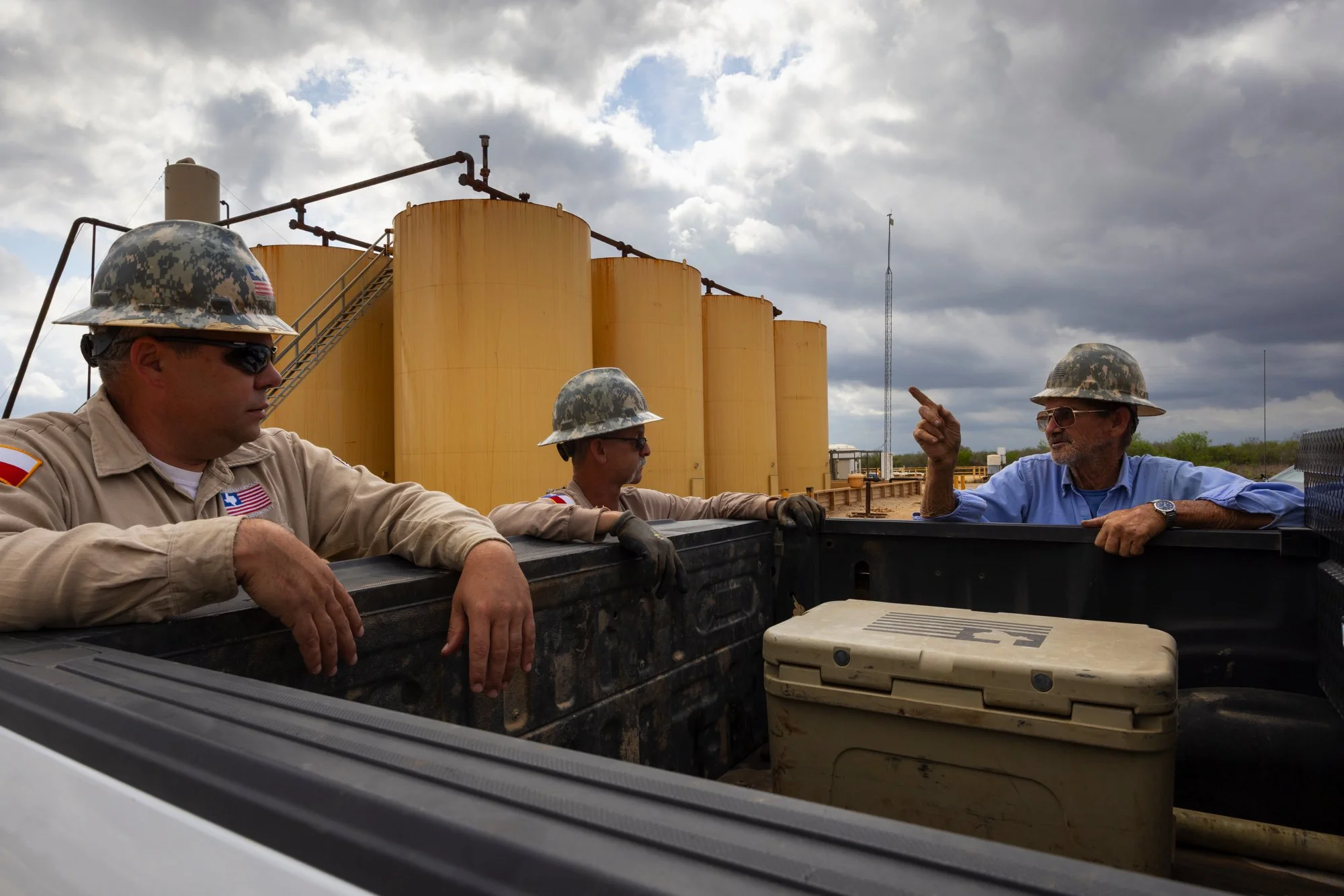 Three men in military uniforms and helmets are sitting on the edge of a truck bed, engaged in a discussion with a man in a blue shirt. Behind them are large yellow tanks and a cloudy sky.