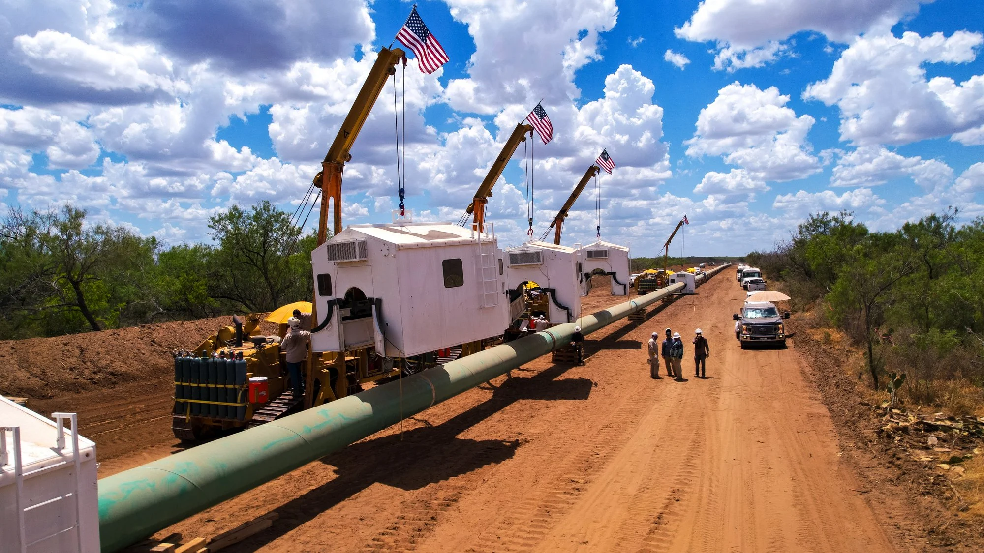 Large pipeline being installed in a desert landscape with a blue sky and scattered clouds. Construction workers and vehicles are present, and there are flags on cranes above the pipeline.