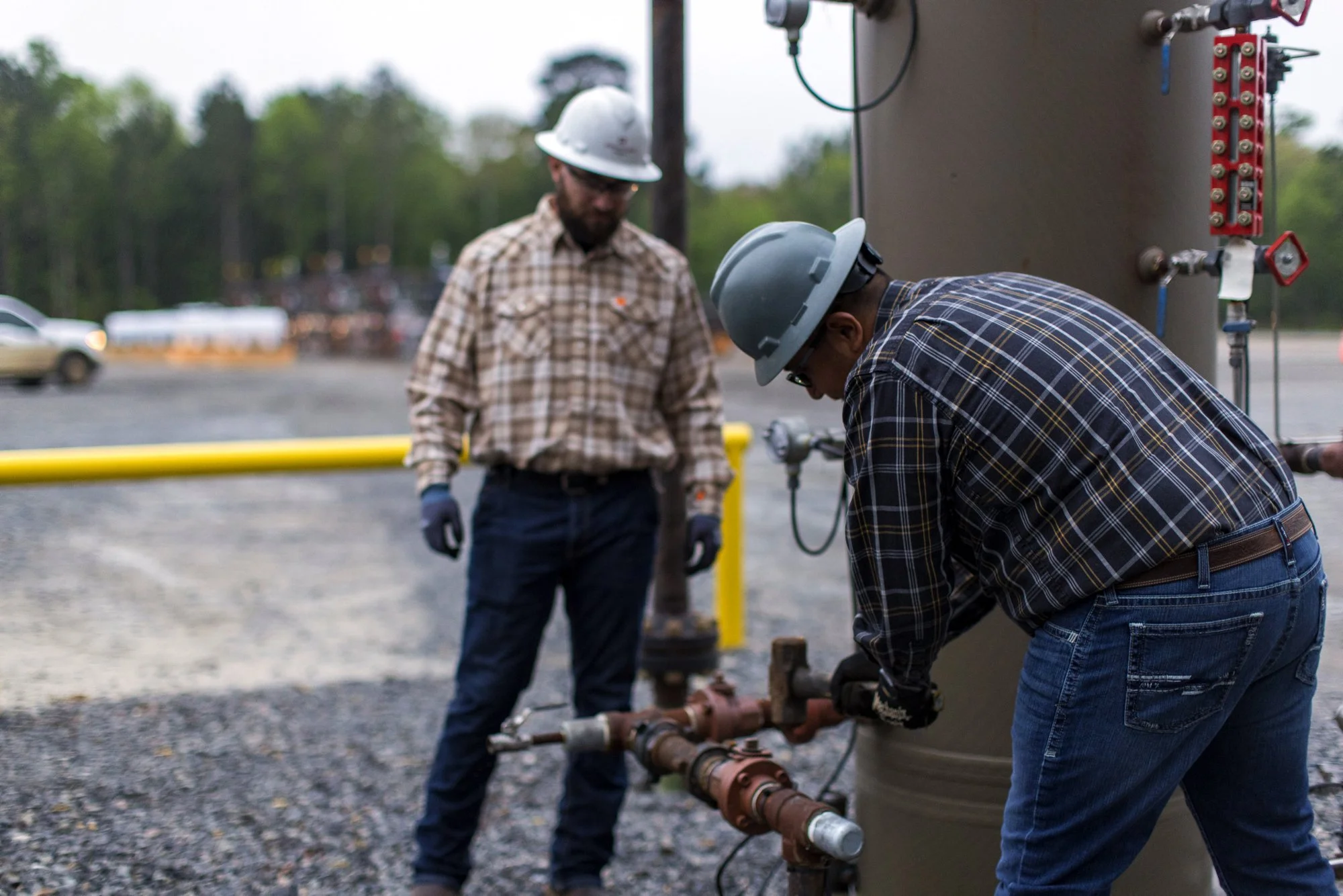 Two workers in safety helmets working on a pipe system outdoors, with trees and vehicles in the background.