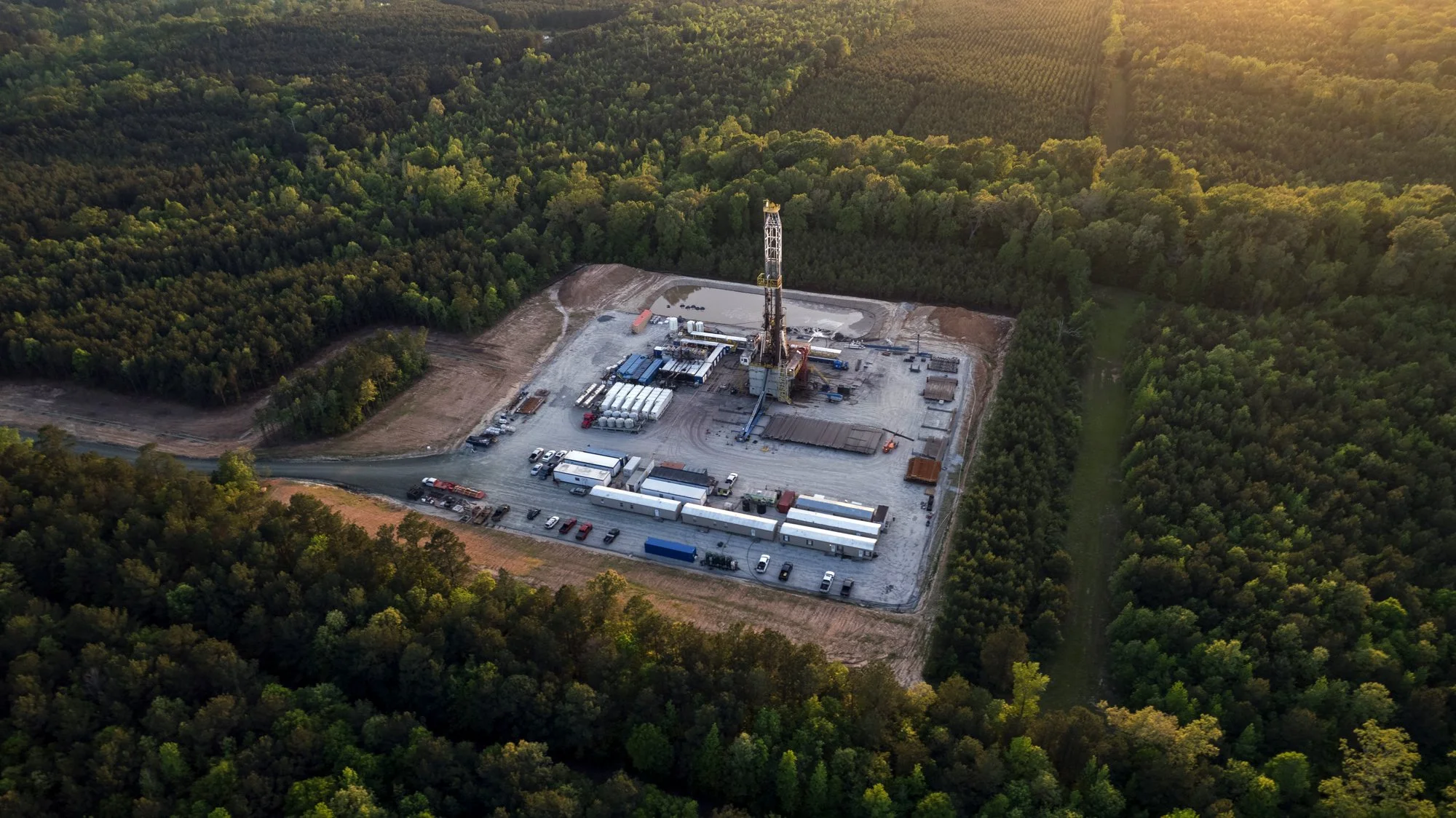 Aerial view of an oil drilling rig in a cleared area surrounded by dense forest, with trucks and equipment nearby.