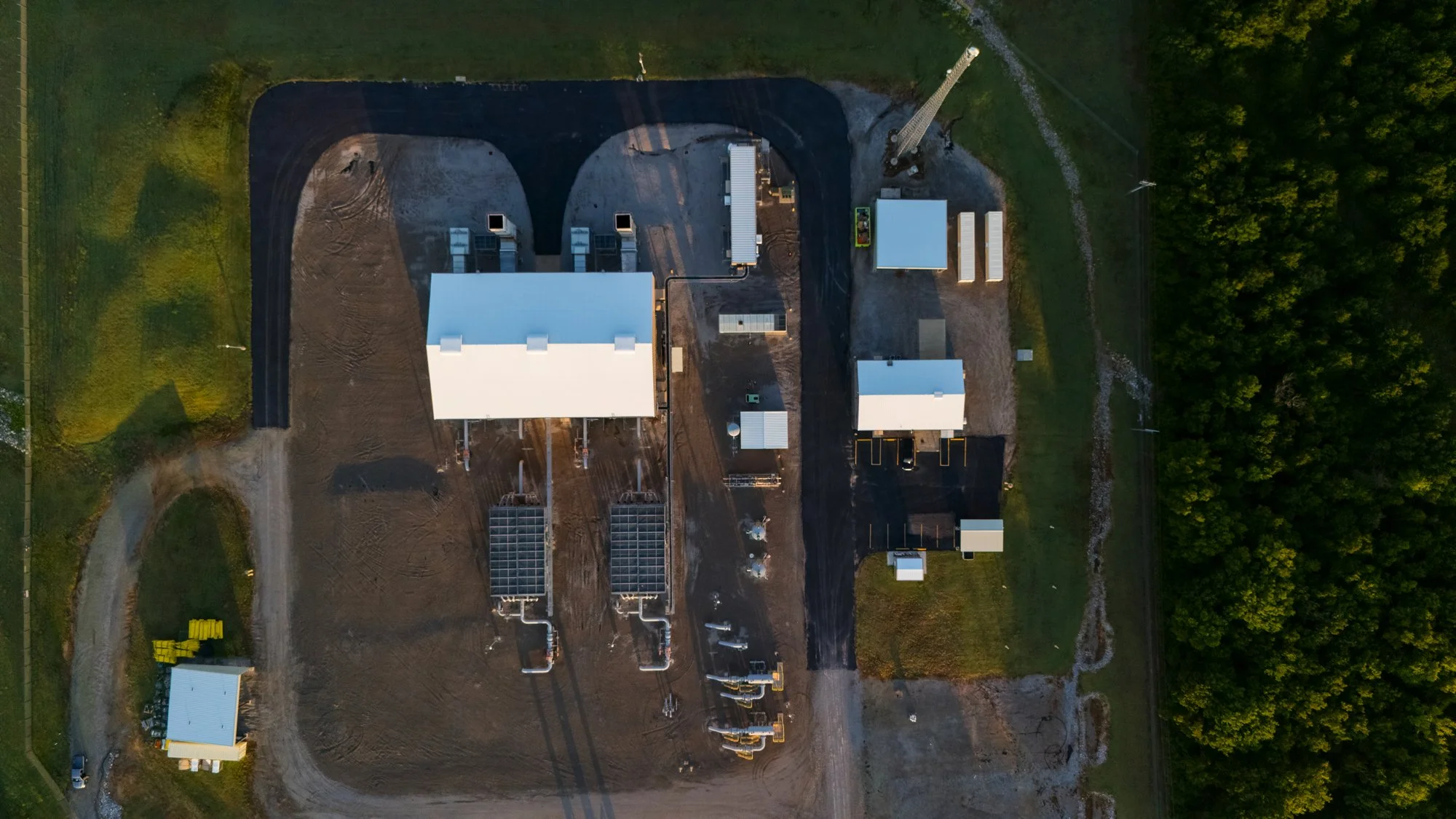 An aerial view of a construction site with buildings, equipment, and a surrounding wooded area.