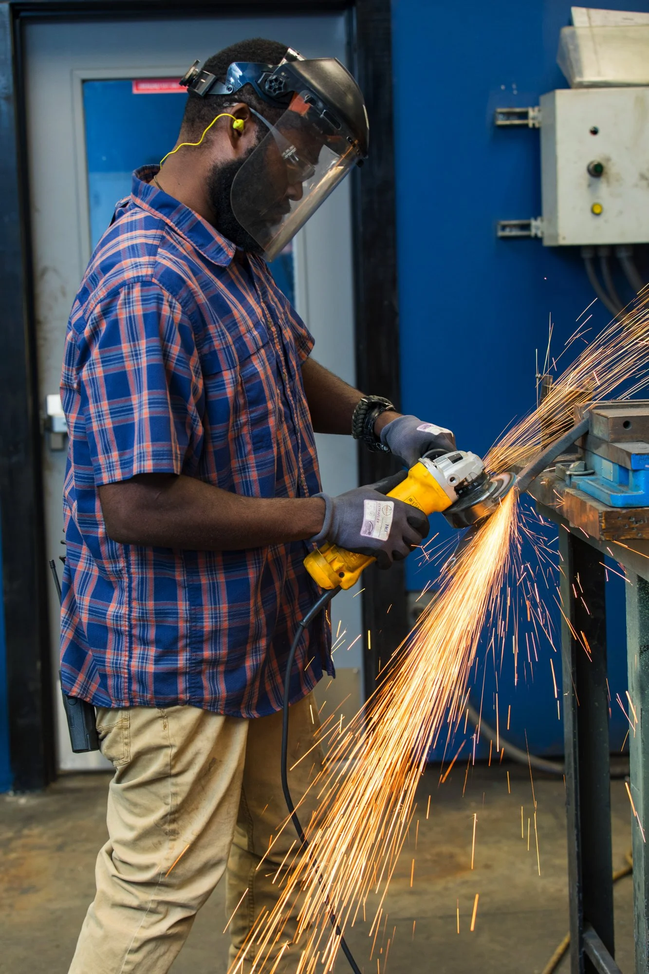 A man wearing safety gear, including a face shield, yellow earplugs, gloves, and casual clothing, uses a power grinder to cut metal, creating sparks in an industrial workshop.