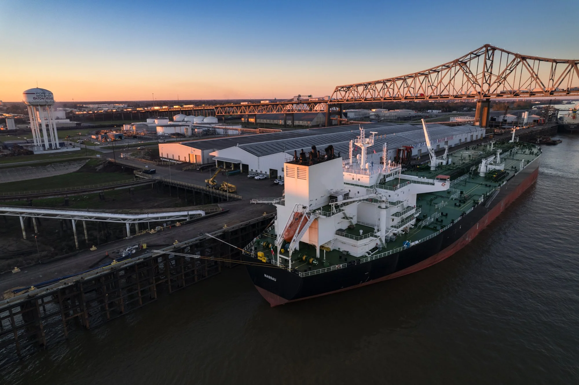 An aerial view of a large cargo ship docked at a port during sunset, with industrial buildings and a bridge in the background.
