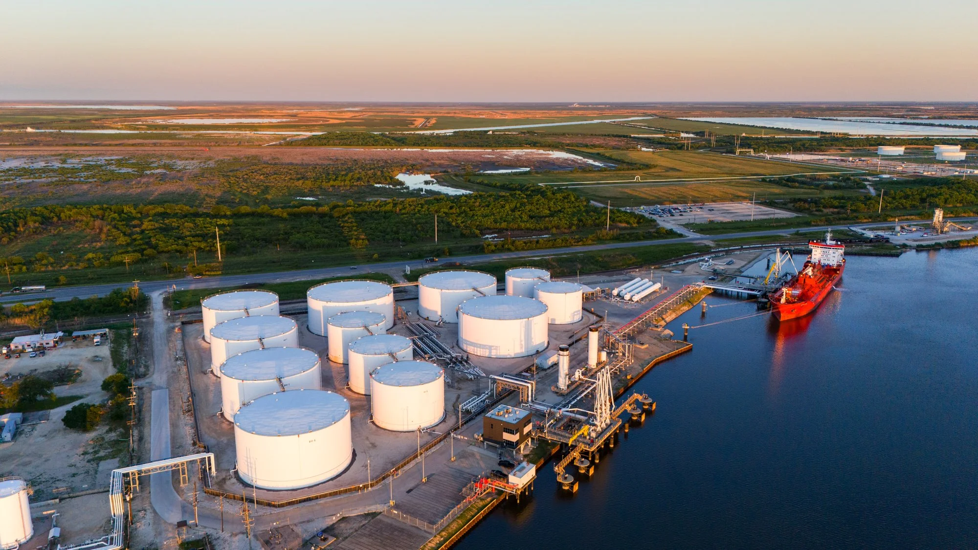 Aerial view of a port with large white storage tanks, a cargo ship docked, and a calm waterway, with green fields and roads in the background.