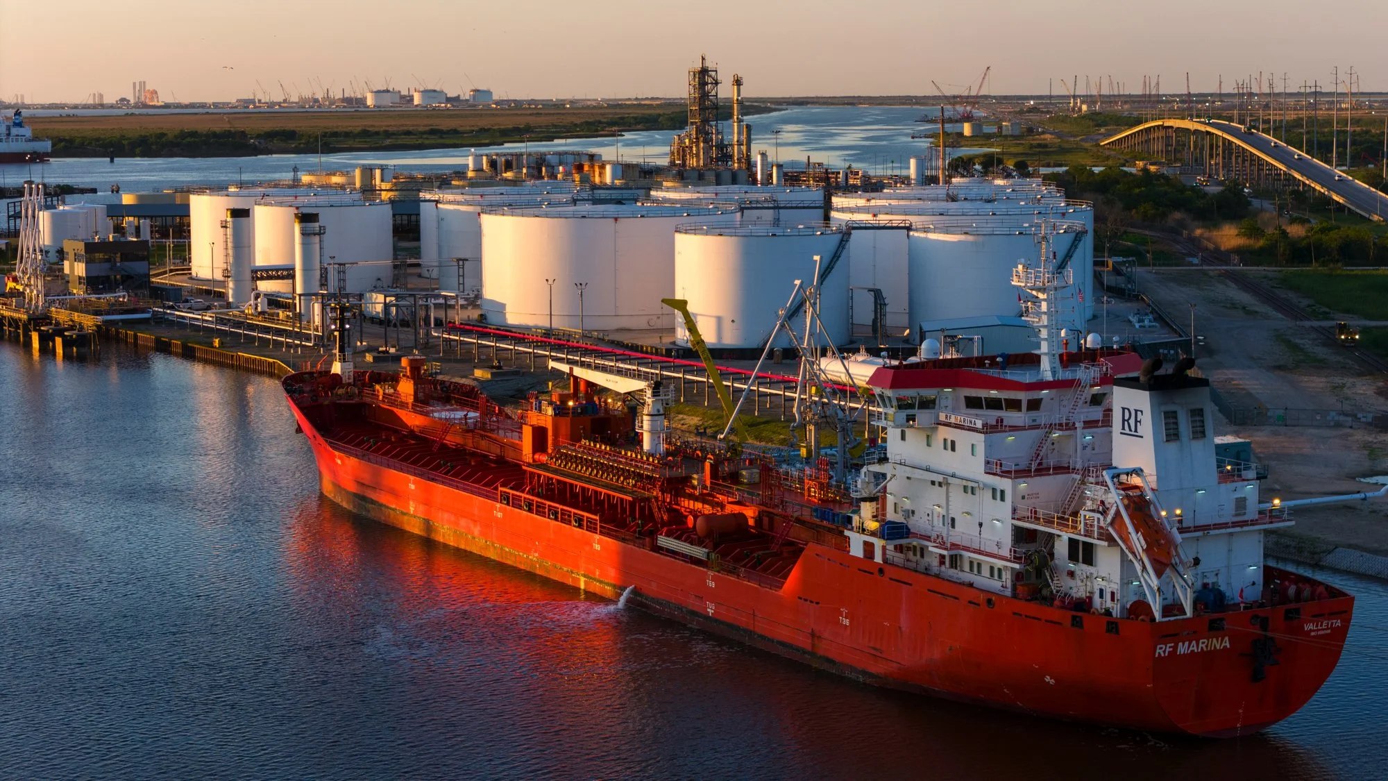 A large red cargo ship, named RF Marina, docked in a port near white storage tanks. The port is surrounded by water, with industrial tanks and cranes in the background, under a sky during sunset.