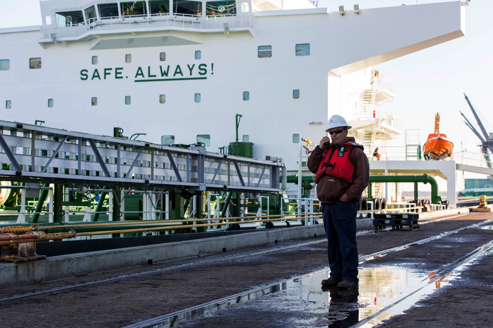 A man standing on a dock talking on a cell phone, wearing a white safety helmet, sunglasses, a red life jacket, and brown jacket. Behind him is a large white ship with the words "SAFE. ALWAYS!" painted on its side. The dock surface is wet with puddle