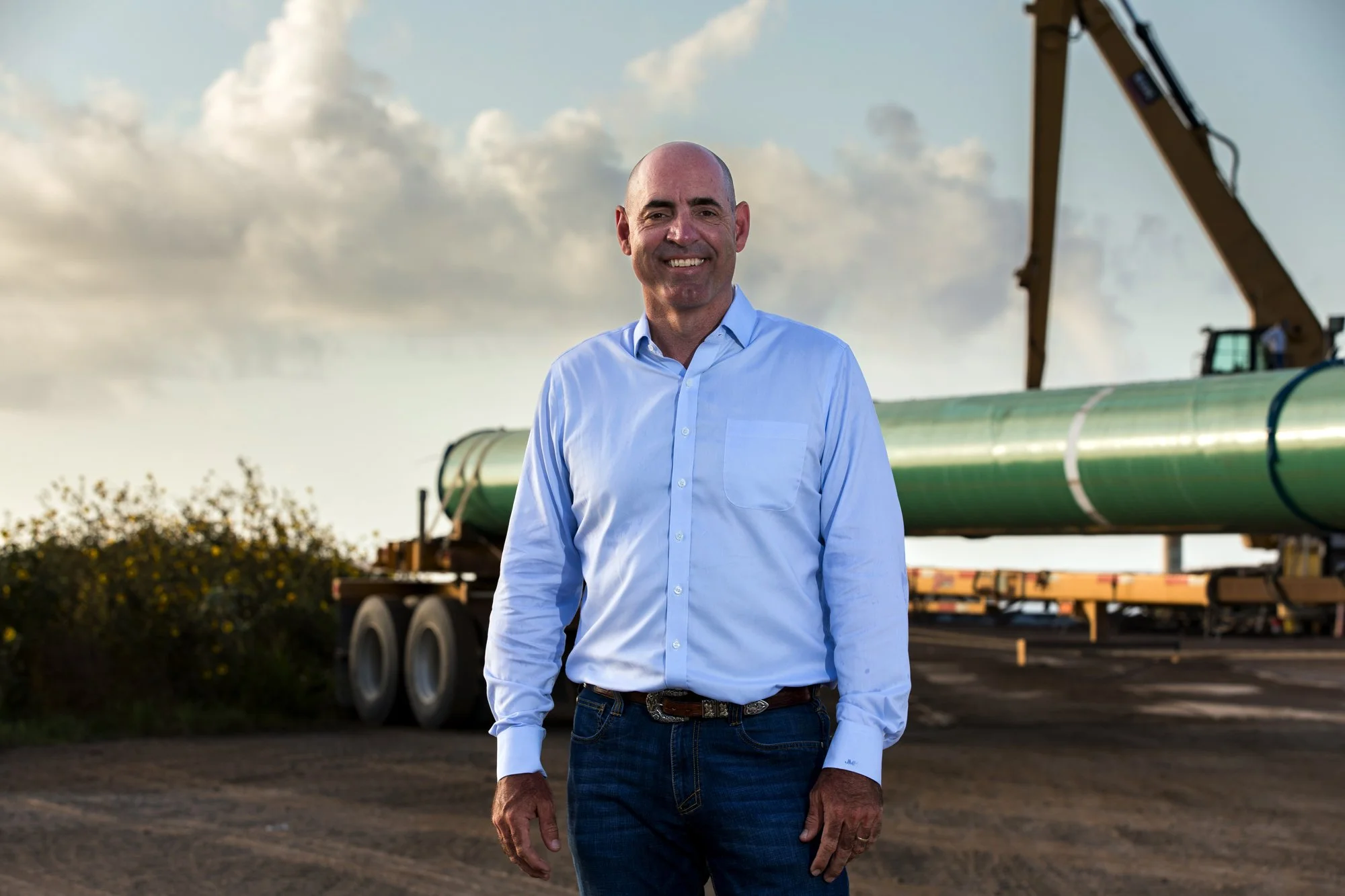 A man in a light blue button-up shirt and jeans standing outdoors near a large industrial pipe and construction equipment, with cloudy sky in the background.