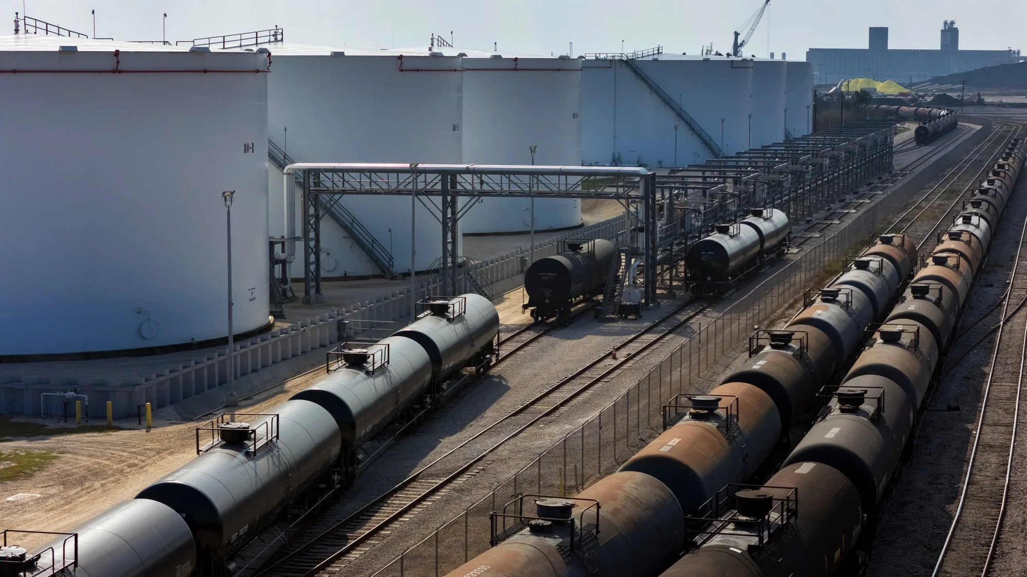 Railway tank cars on tracks near fuel storage tanks with industrial structures in the background.