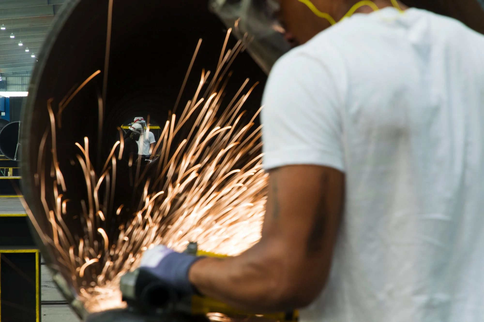 A worker in a white shirt and gloves is welding a large metal pipe with sparks flying. Two workers in the background are wearing protective gear, including a helmet with a face shield, in an industrial setting.