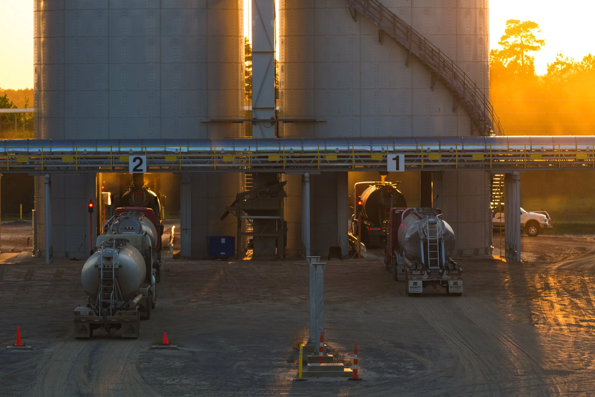 Two cement trucks parked under silos at a construction site during sunset.