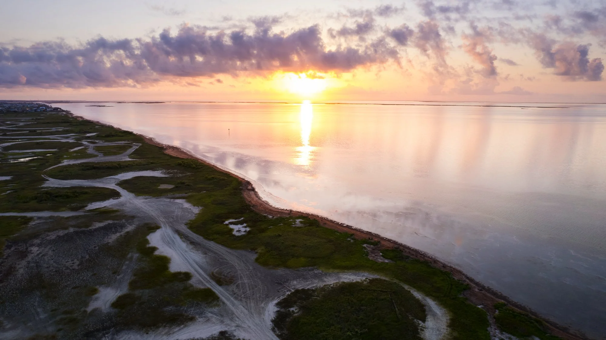 A sunset over a calm body of water with pink, purple, and orange clouds in the sky, reflecting on the water's surface. A narrow strip of land with winding dirt paths and patches of green vegetation runs along the left side of the image.