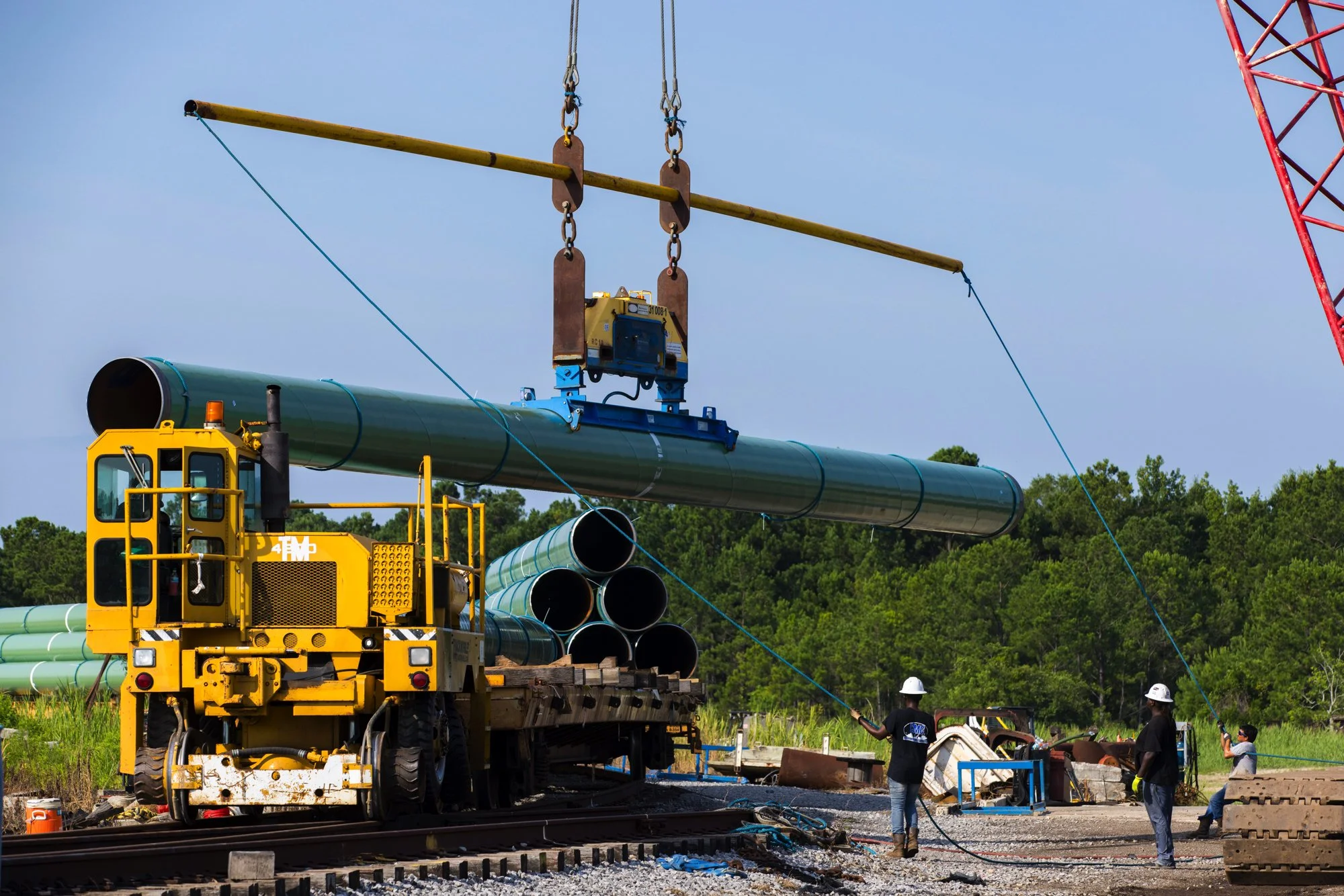 Workers operate a crane to install large green pipes at a construction site next to railway tracks, with trees in the background.