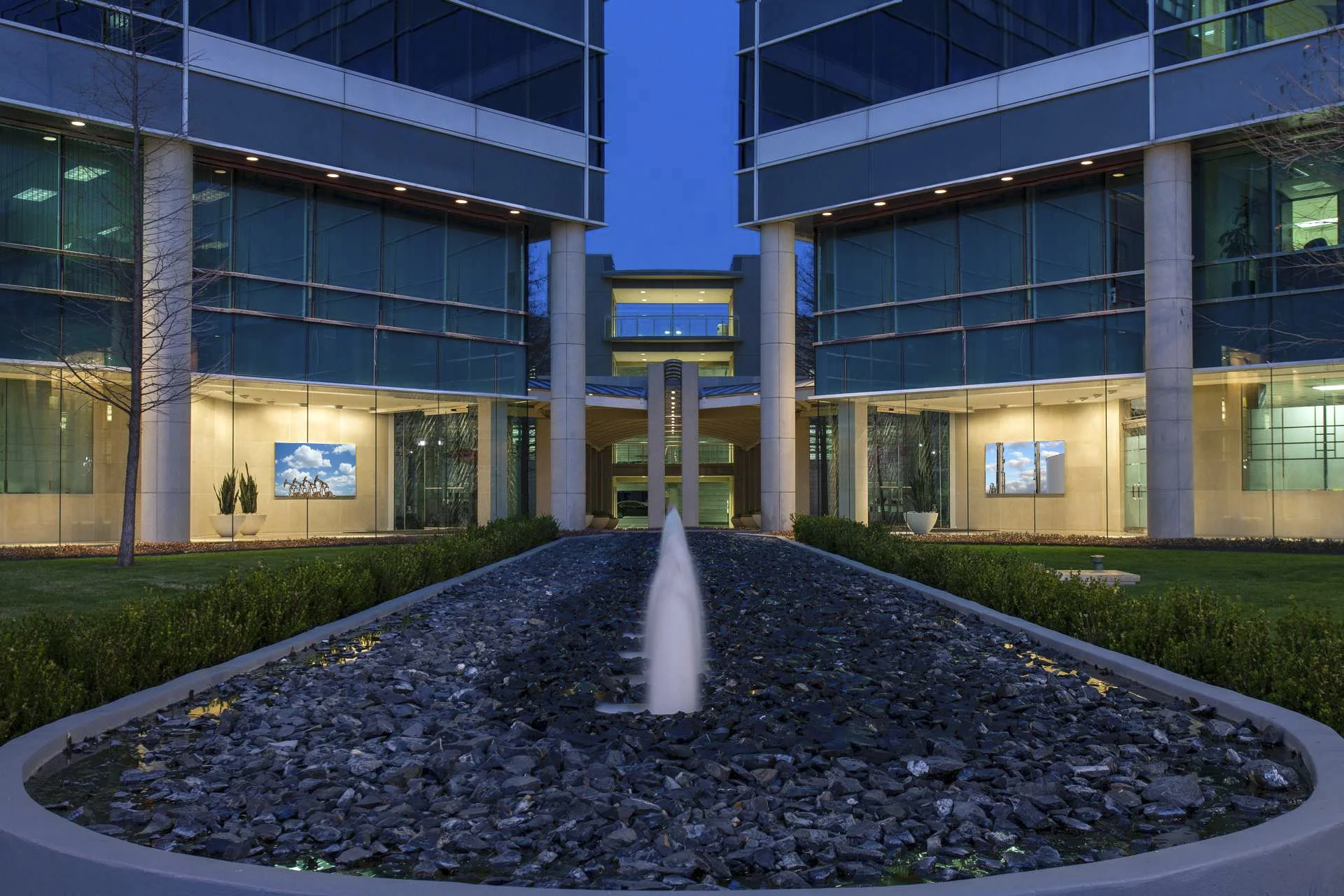 Modern office building with glass facade, illuminated windows, and a central courtyard featuring a water fountain, trees, and landscaped greenery at dusk.