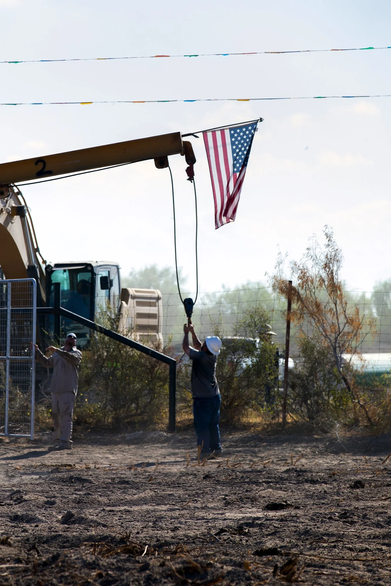 Two workers with helmets installing an American flag on a flagpole at a construction site with dusty ground and trees in the background.