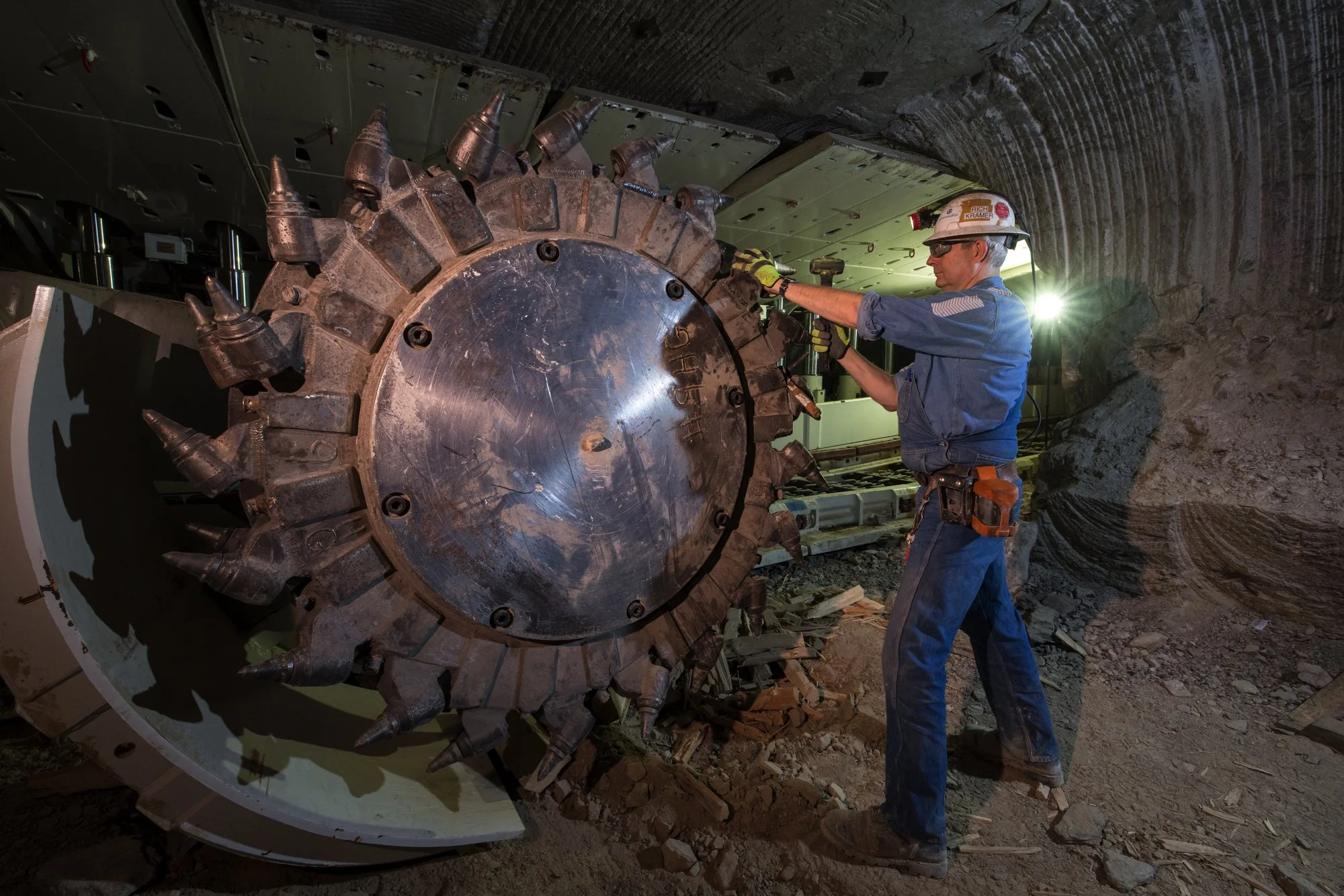 A construction worker wearing a safety helmet, glasses, and a blue uniform is working on a large tunnel boring machine inside a tunnel.