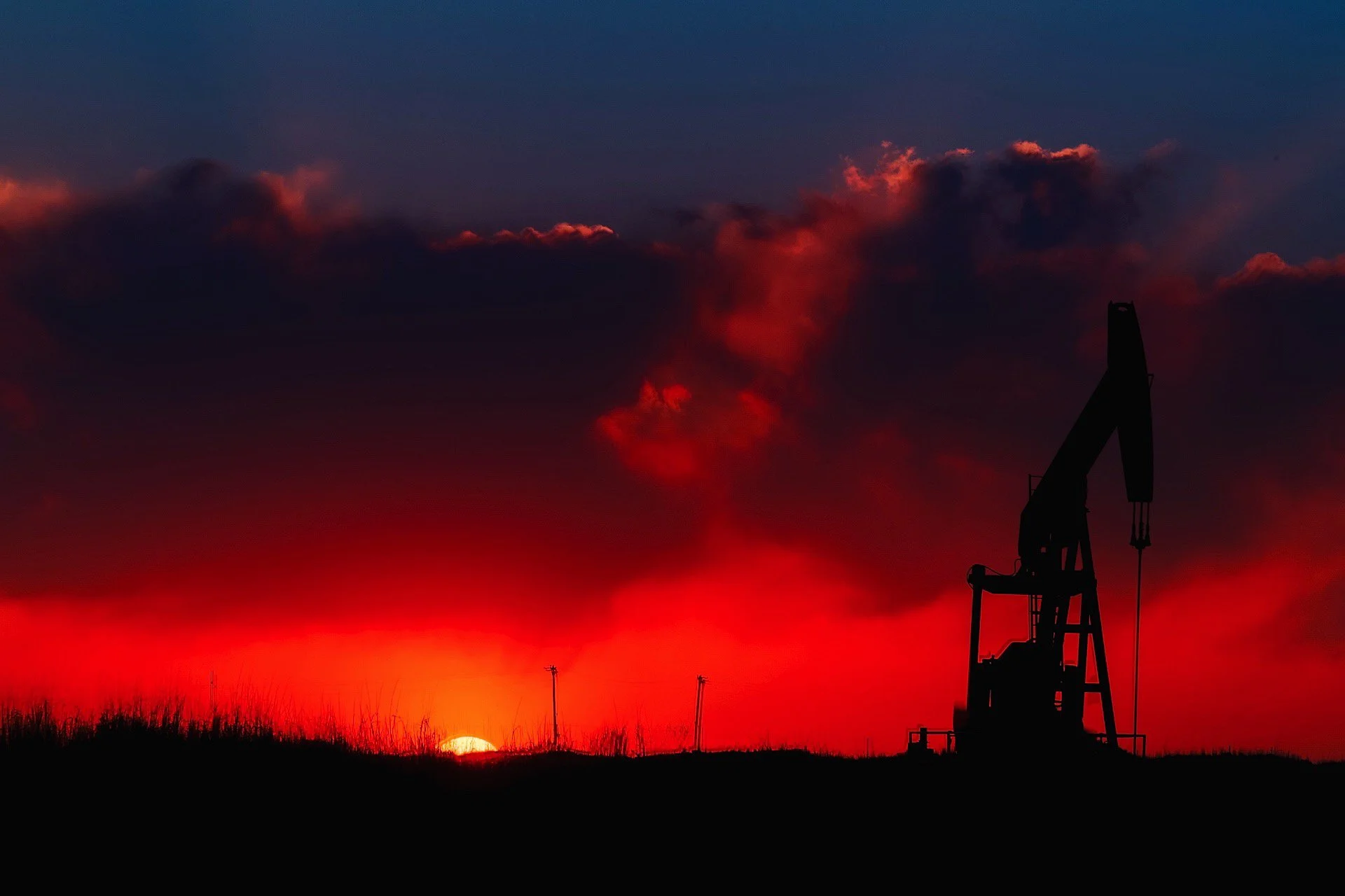 Silhouette of an oil pumpjack against a vivid red and orange sunset sky with clouds.