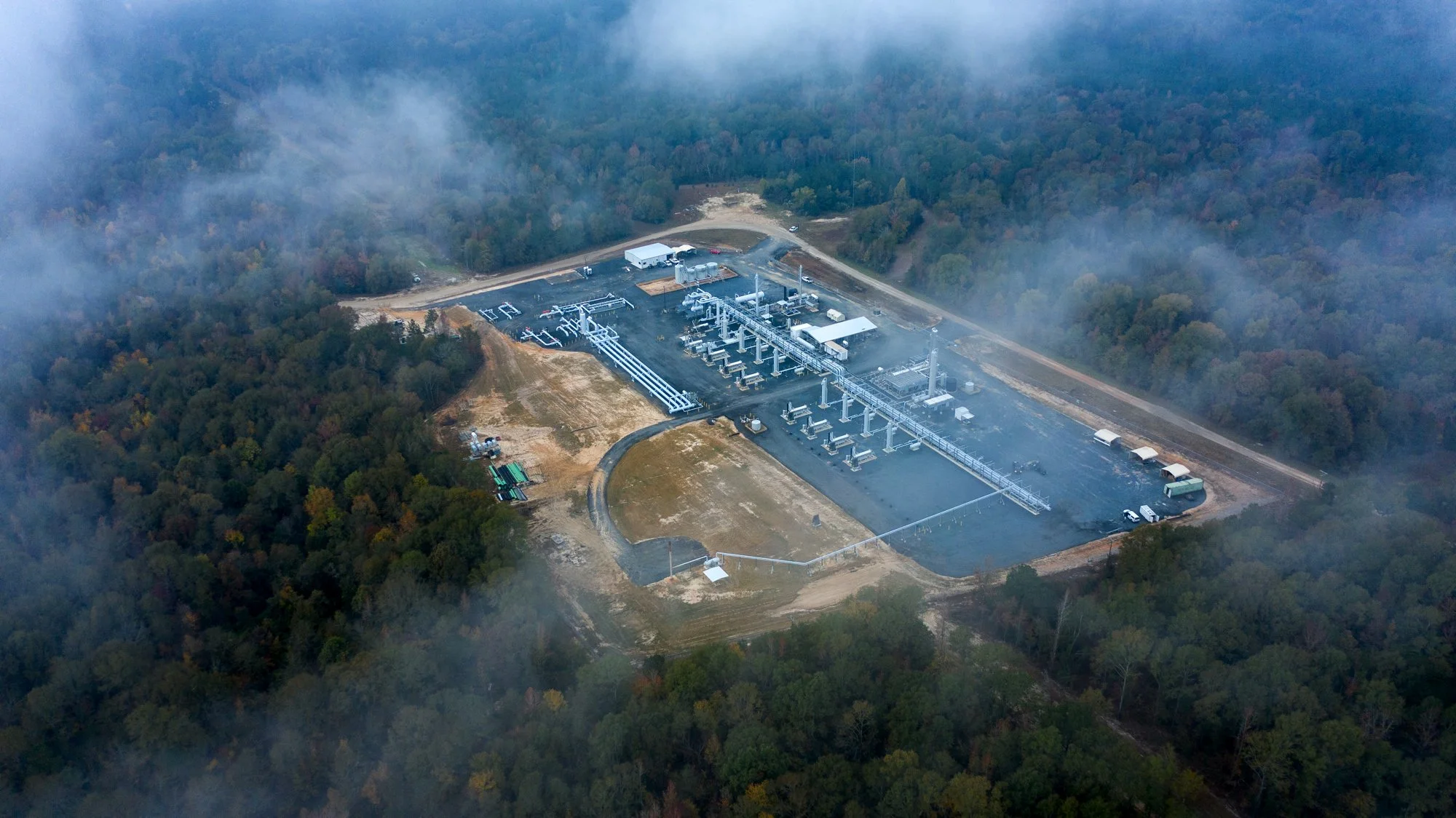 Aerial view of an industrial facility, possibly a water treatment or power plant, surrounded by dense forest and fog.