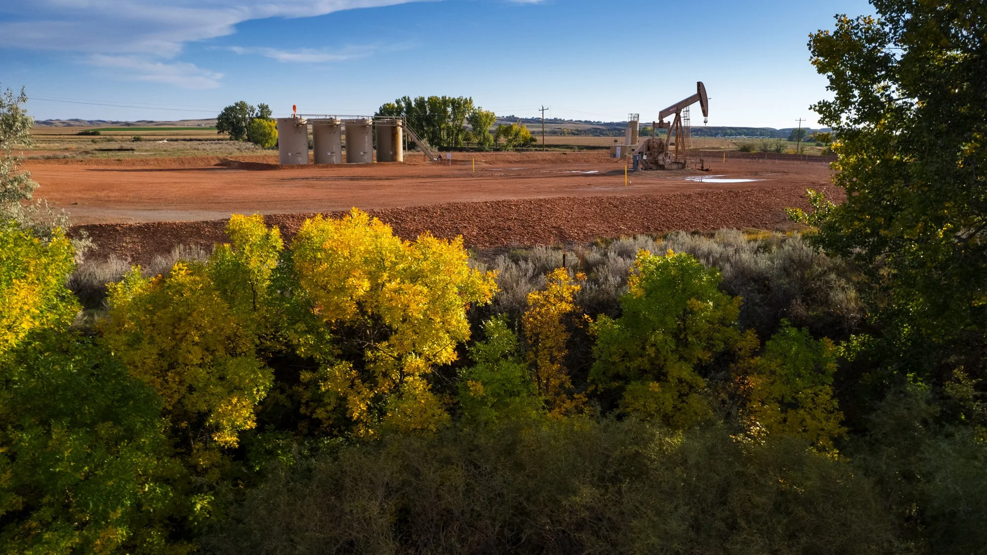Oil pumpjack and storage tanks on red dirt land, with trees displaying fall colors in the foreground and a flat landscape under a blue sky.