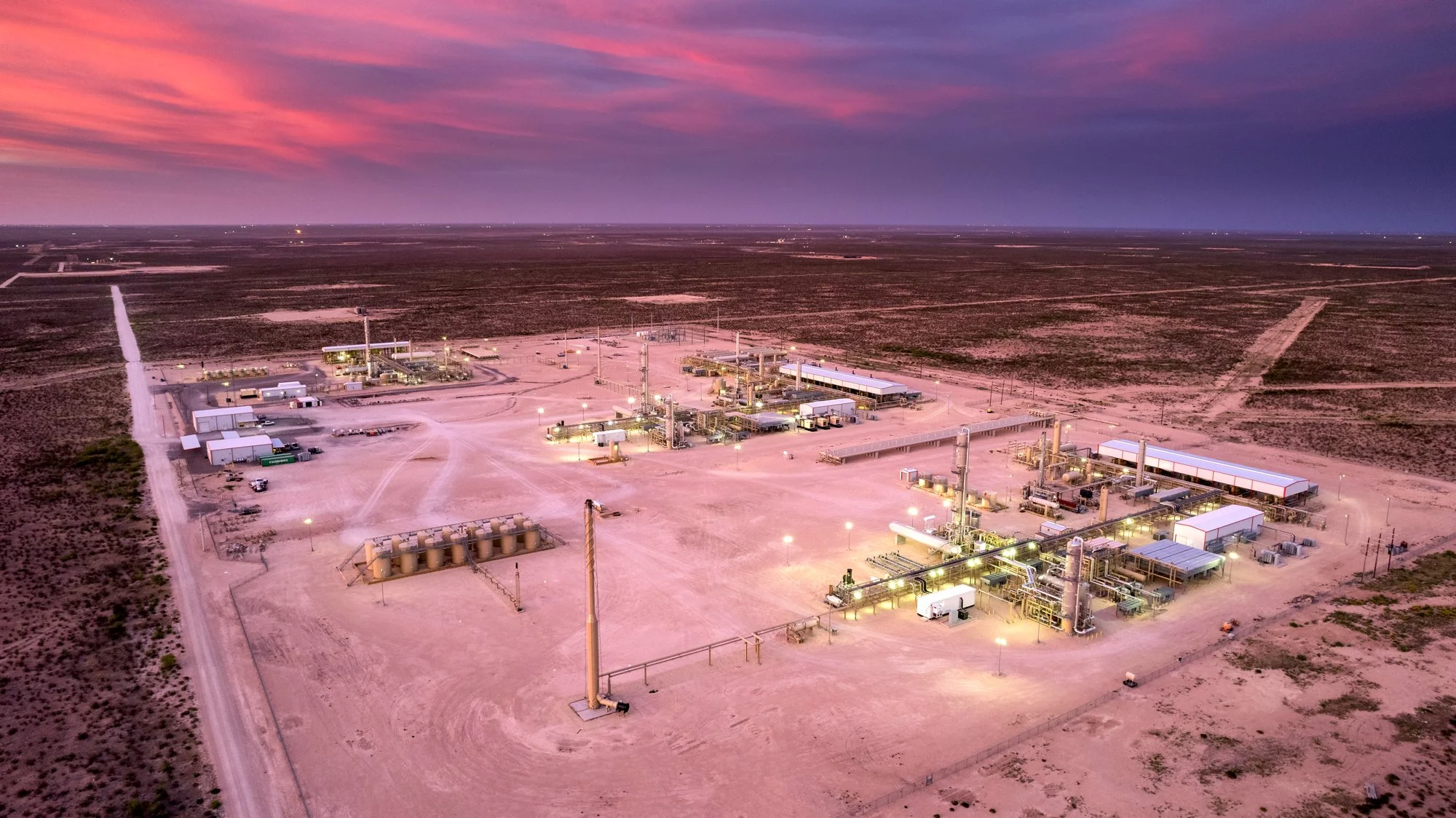 An aerial view of an oil or gas processing facility in a desert, with multiple pipelines, storage tanks, and structures illuminated at dusk, surrounded by flat, arid land with little vegetation.