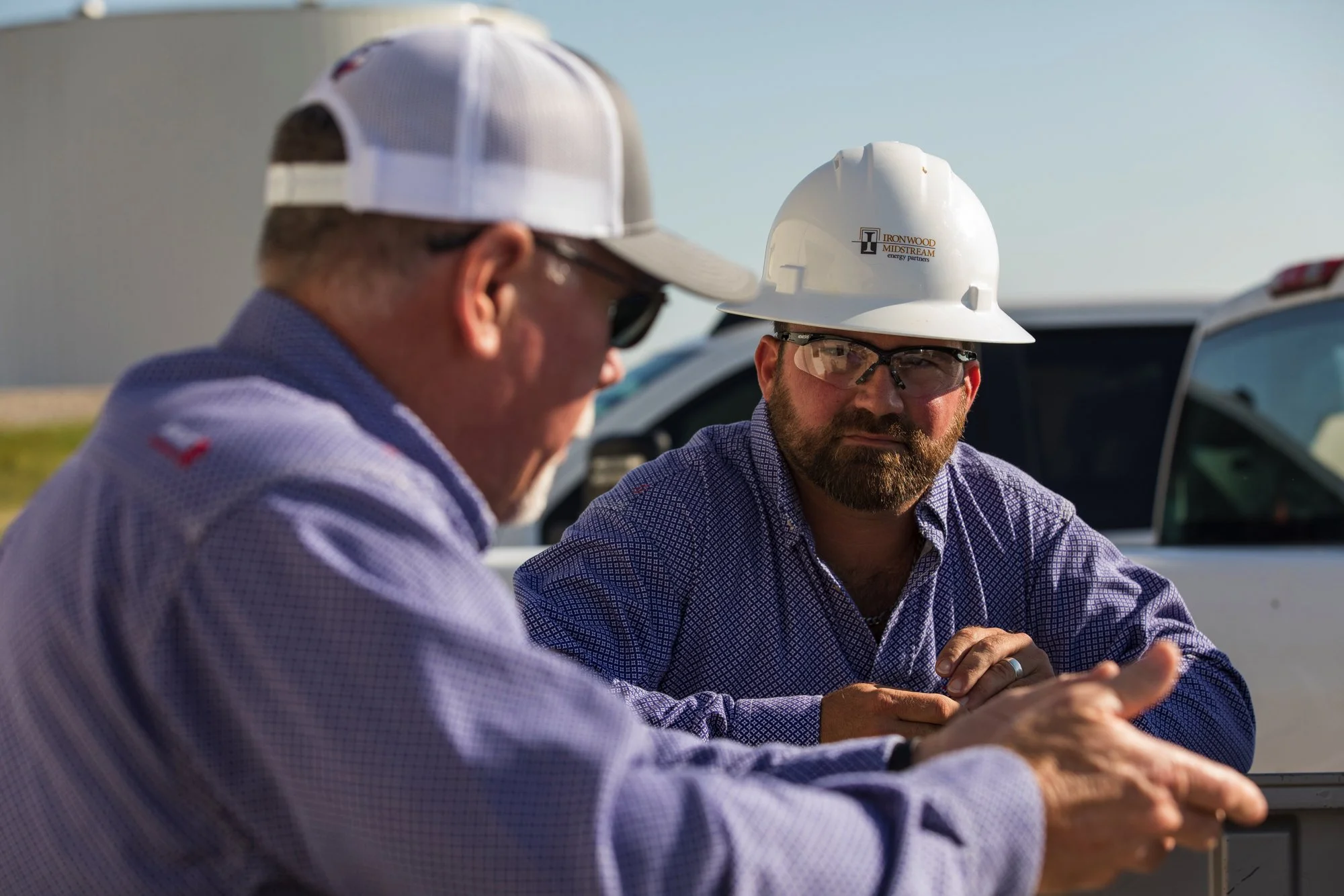 Two construction workers talking outdoors, one wearing a white hard hat with a company logo, safety glasses, and a blue checkered shirt, the other wearing a white cap, sunglasses, and a blue shirt, with cars and a building in the background.